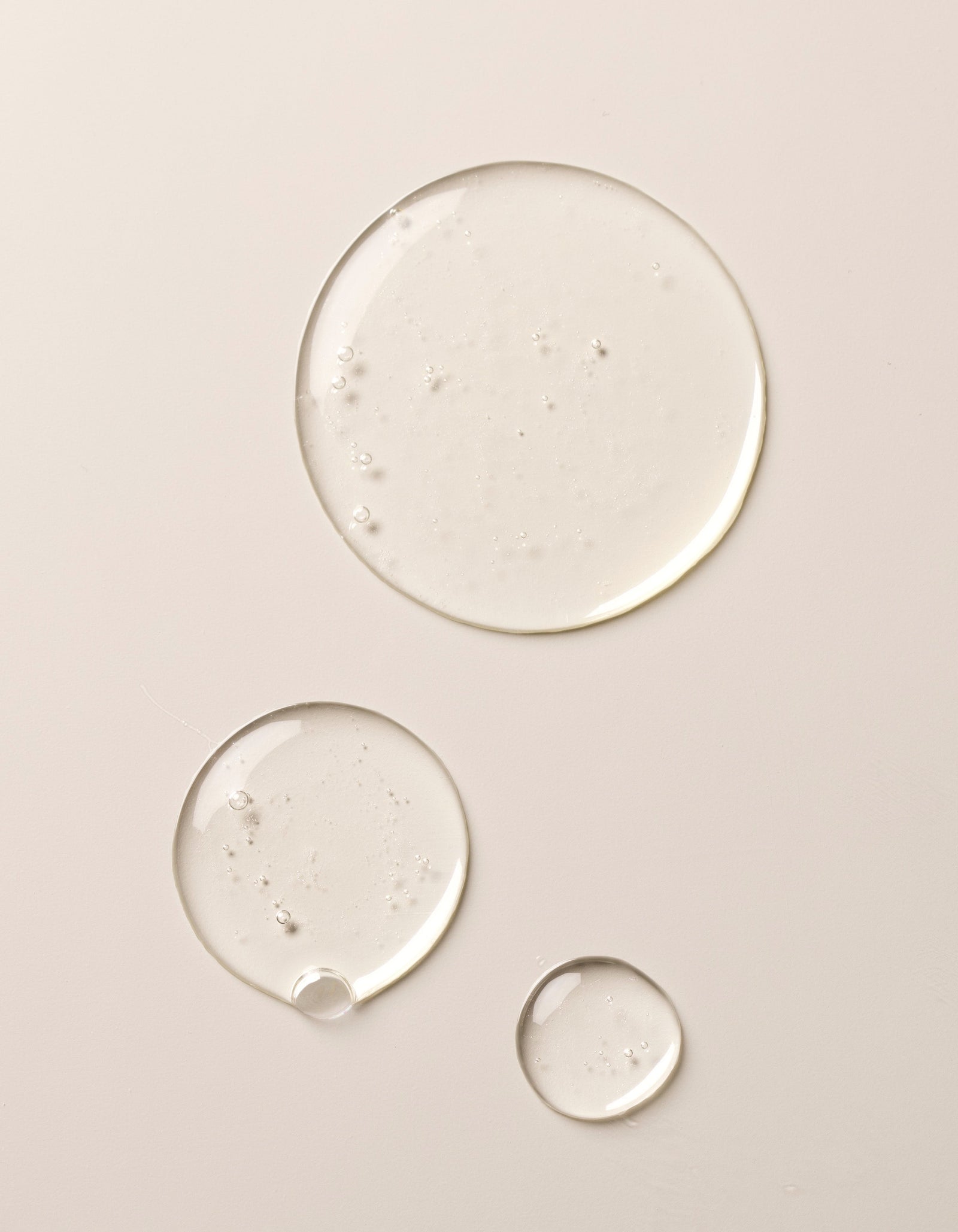Three glossy, clear droplets of Cozy Earth's Double Cleanse Duo rest on a smooth, light beige surface. The reflective liquid forms vary in size and contain small visible air bubbles.