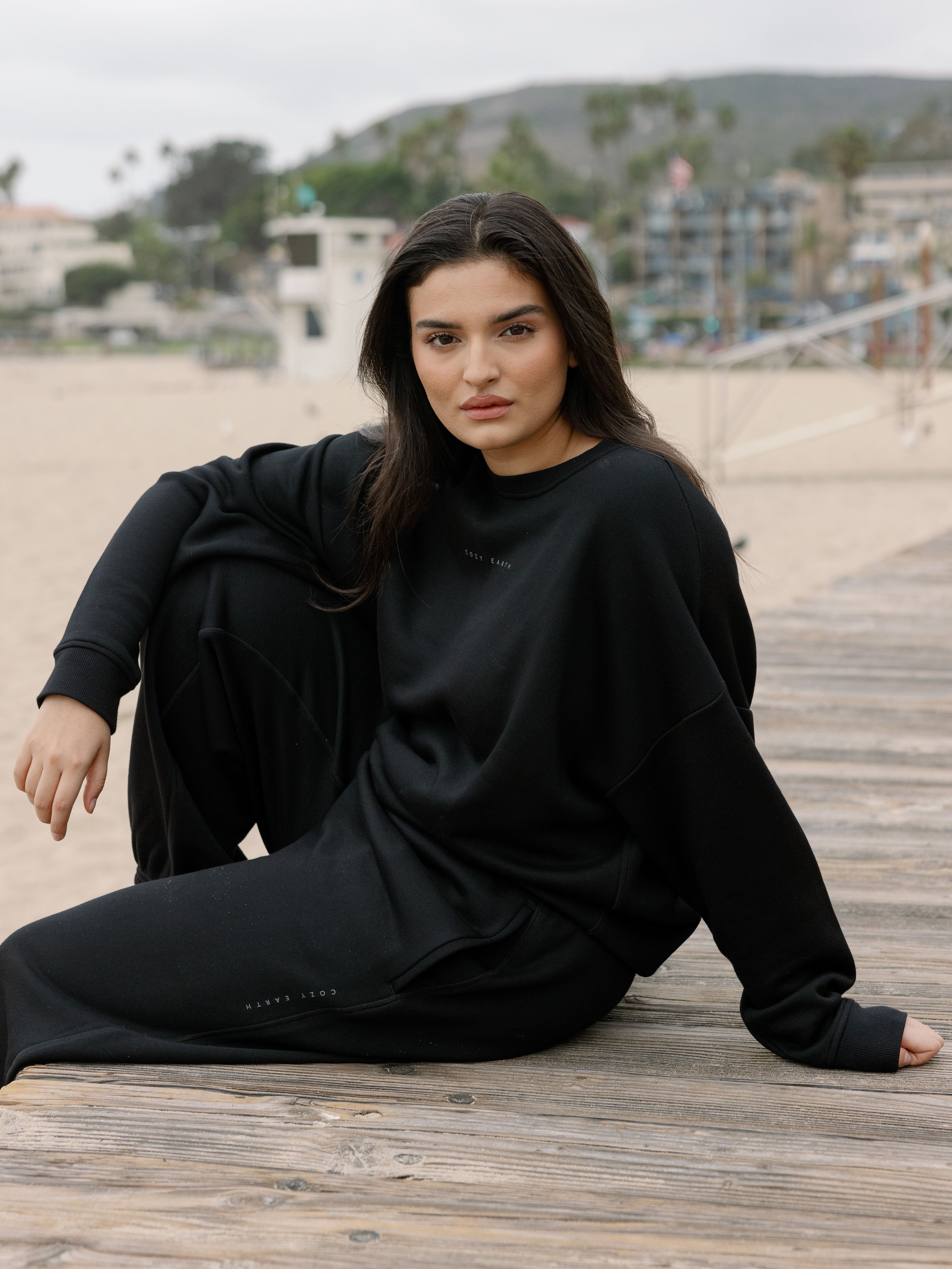 Black CityScape Pullover Crew. The Pullover is being worn by a female model. Accompanying city scape clothing is being worn to complete the look of the outfit. The photo was taken with a sandy beach background. 