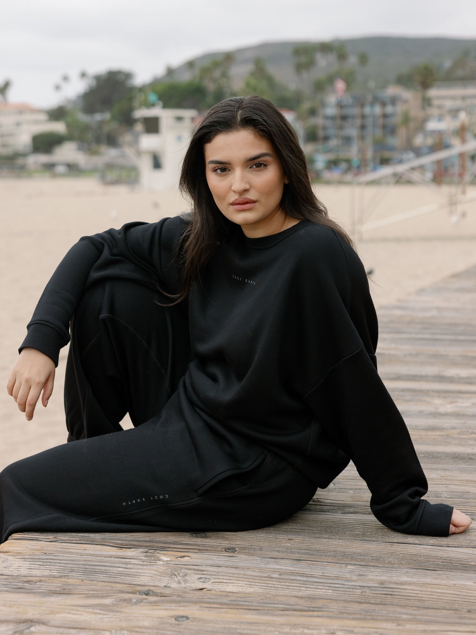 Black CityScape Pullover Crew. The Pullover is being worn by a female model. Accompanying city scape clothing is being worn to complete the look of the outfit. The photo was taken with a sandy beach background. 
