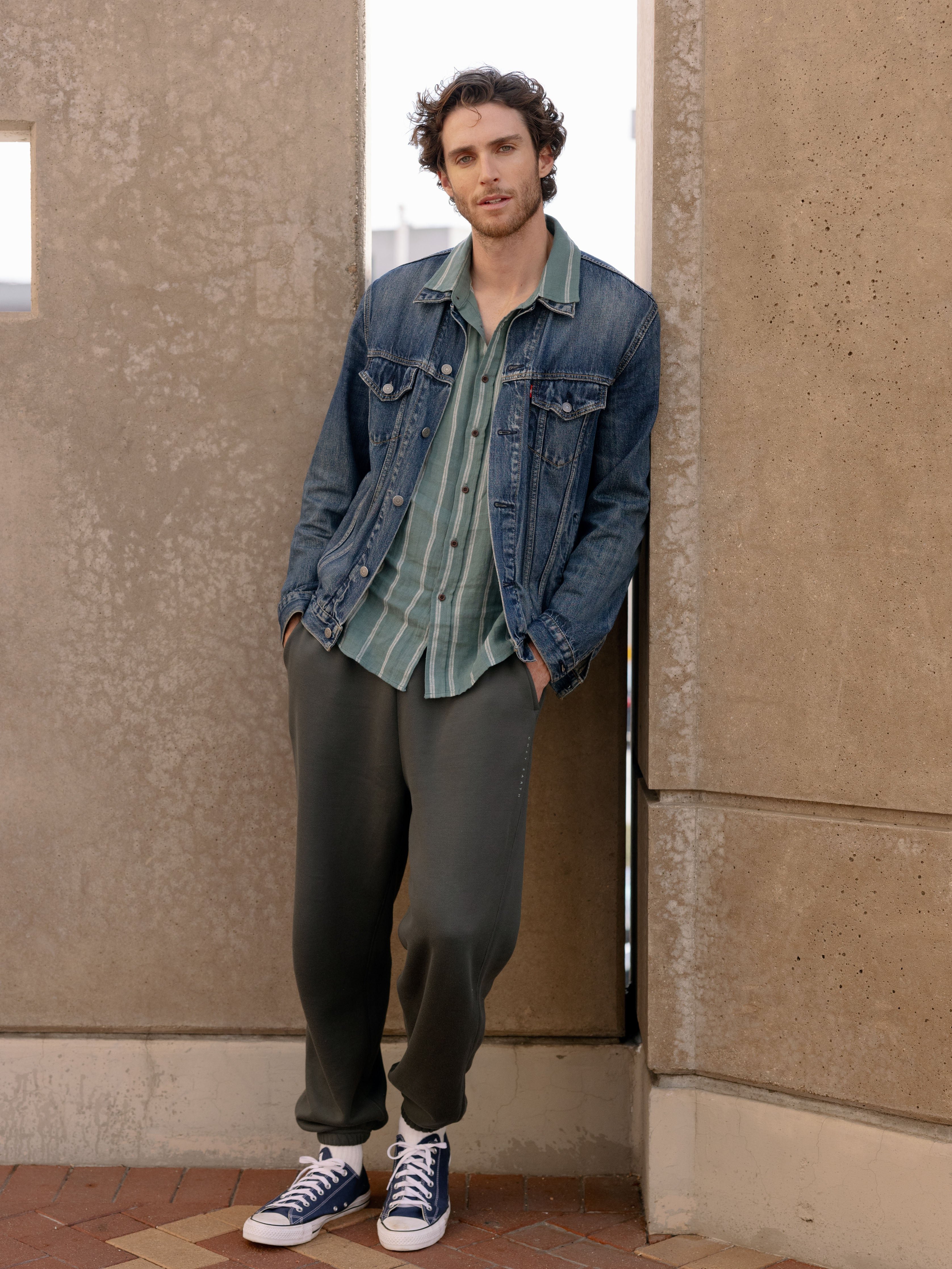 Man wearing storm cityscape sweats and denim jacket next to city wall 