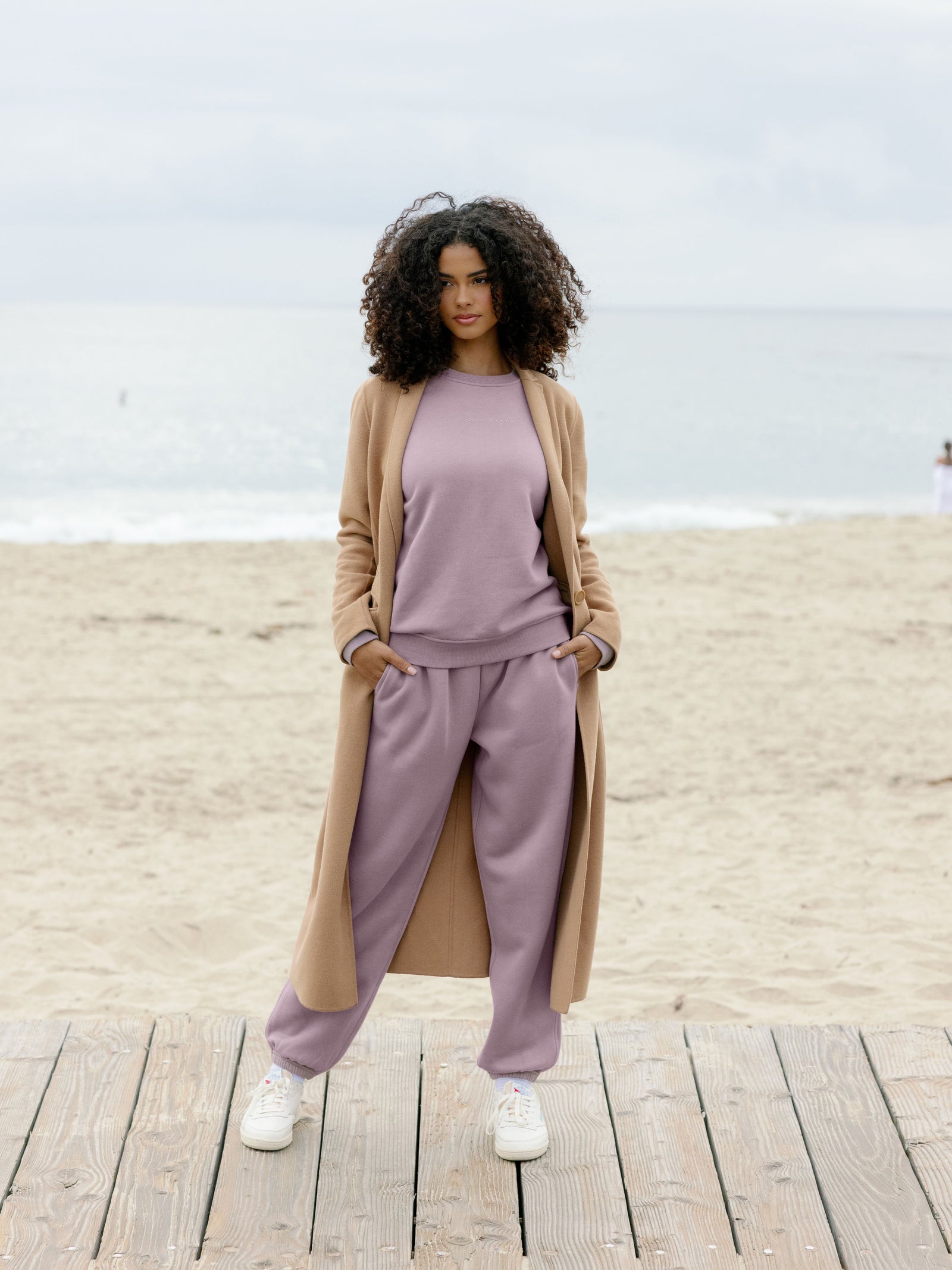 Dusty Orchid CityScape Pullover Crew. The Pullover is being worn by a female model. Accompanying city scape clothing is being worn to complete the look of the outfit. The photo was taken with a sandy beach background. 
