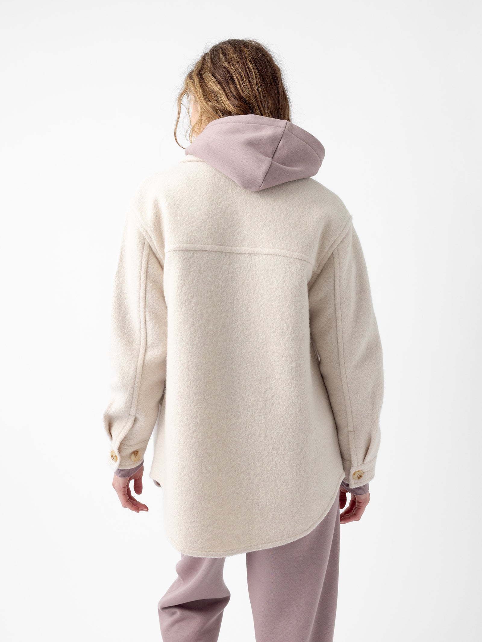 A person stands with their back to the camera, wearing the Cozy Earth Women's Boucle Shacket, an oversized light cream wool jacket featuring brown buttons and a pink hood. They are also dressed in matching pink pants, with a plain white backdrop in the background.  