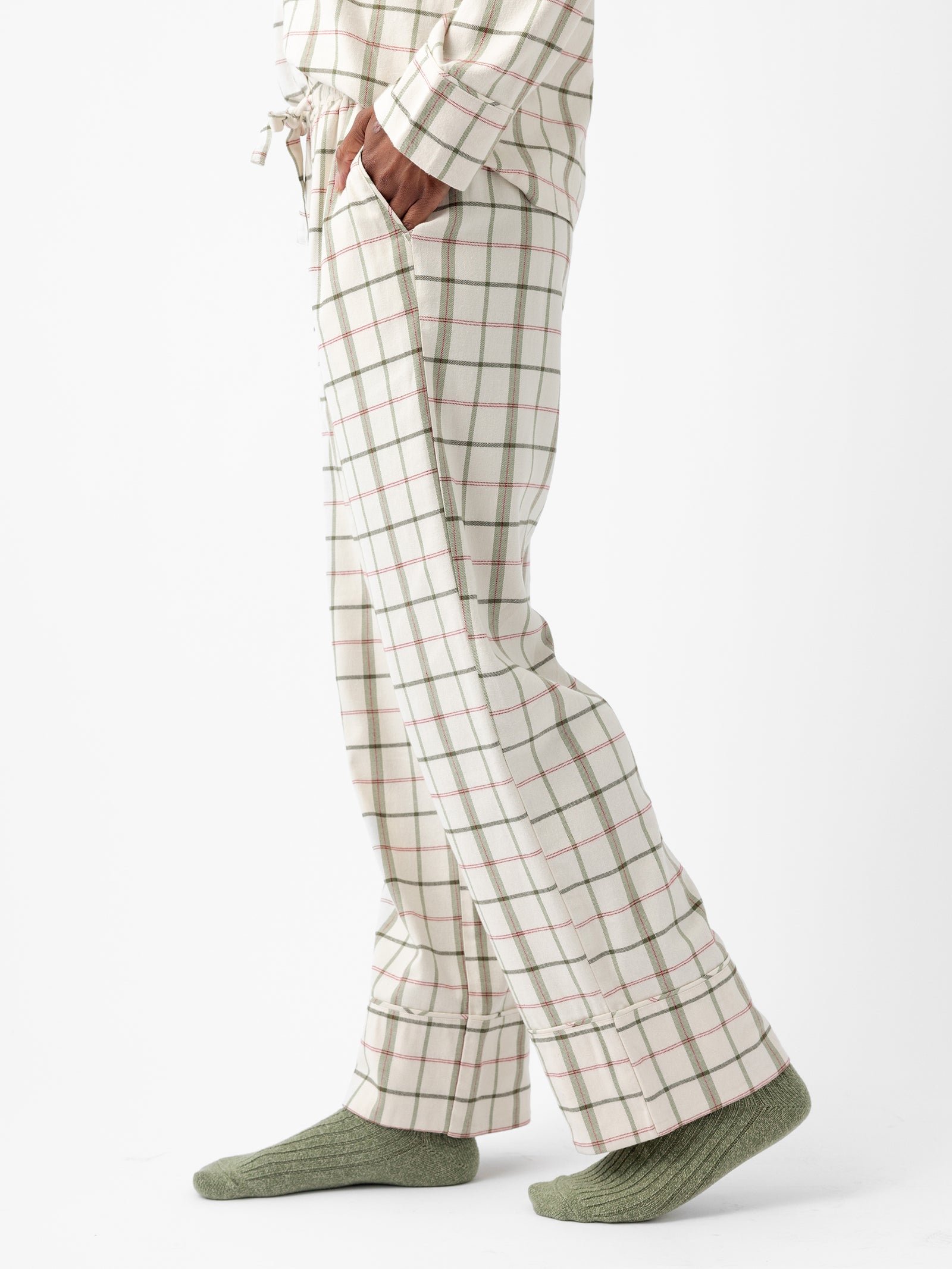 A person wearing the Women's Farmhouse Long Sleeve Pajama Set by Cozy Earth stands on a white background. The pajamas feature a white base with green and red stripes. The individual has one hand in their pants pocket and is wearing light green socks. Their slightly visible torso is also clad in the matching plaid fabric from the set. 