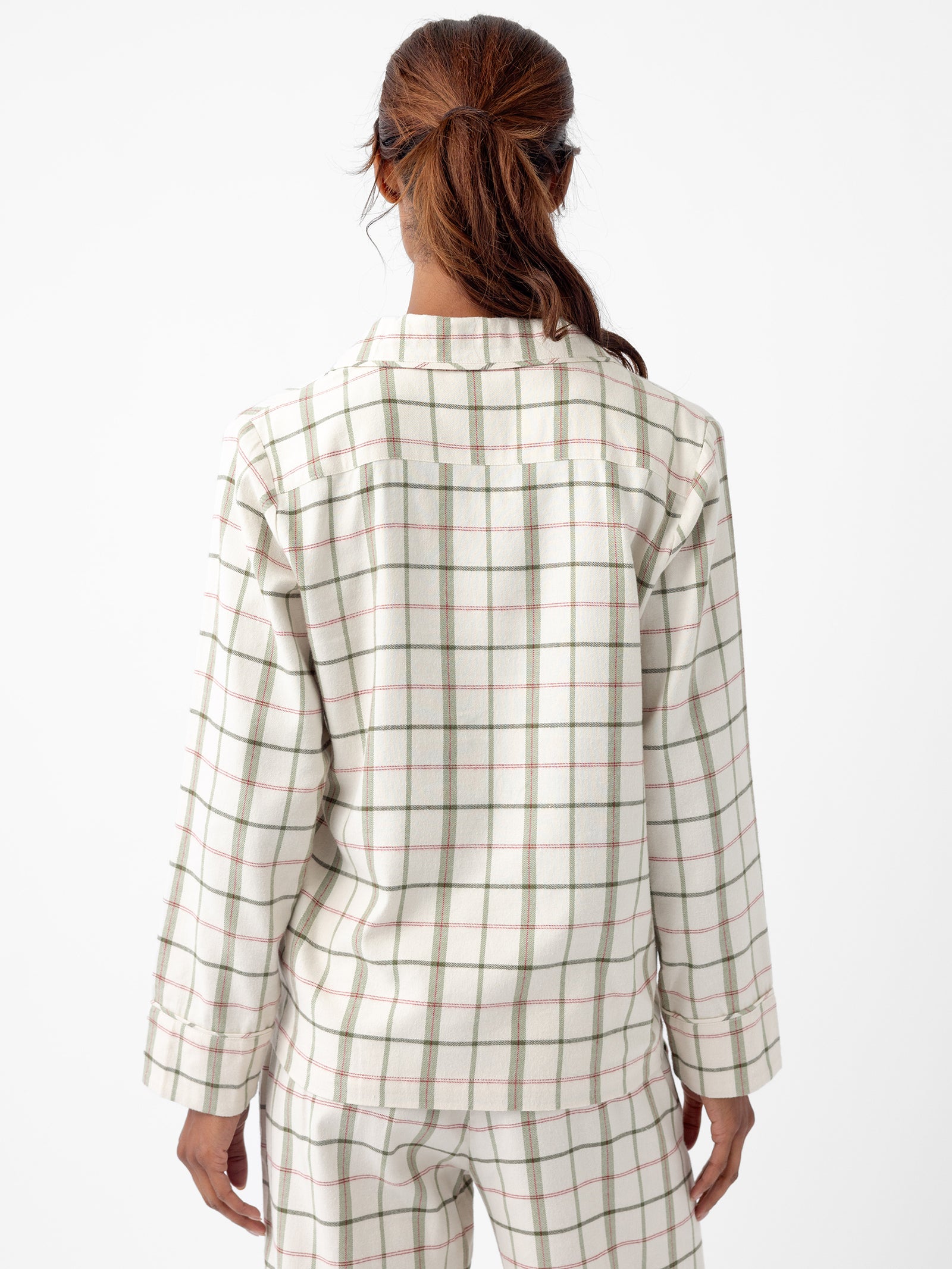 A person with reddish-brown hair tied in a ponytail is seen from behind, wearing the Women's Farmhouse Long Sleeve Pajama Set by Cozy Earth. The pajama set has a white plaid design with green and red stripes, and the background is plain white. 