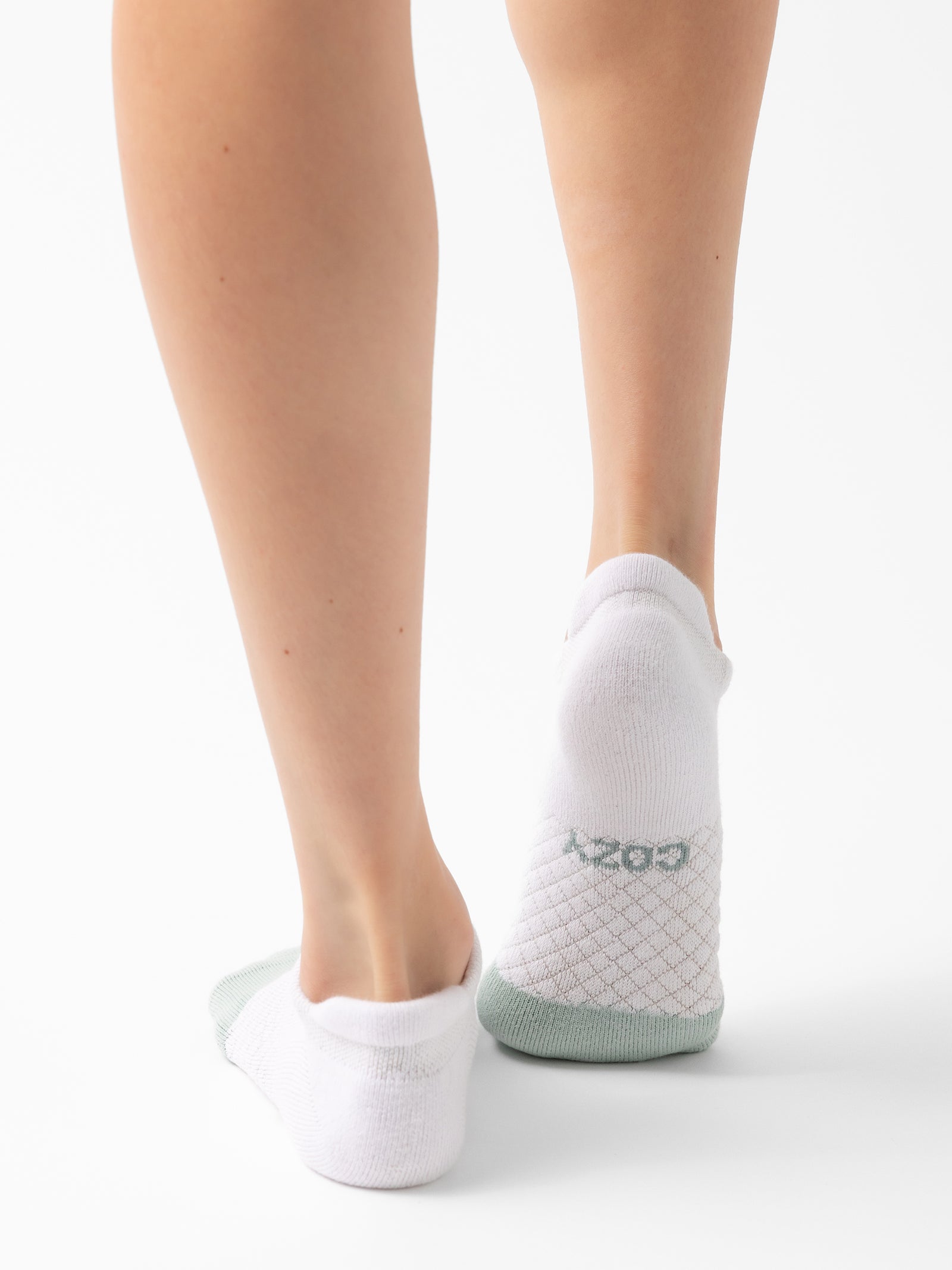 A close-up view captures a person from the calf down wearing white Essential Ankle Socks by Cozy Earth, featuring a light green heel. These socks display a subtle diamond pattern on the sole and have "Bombas" written in green on the back. The background is plain white. 