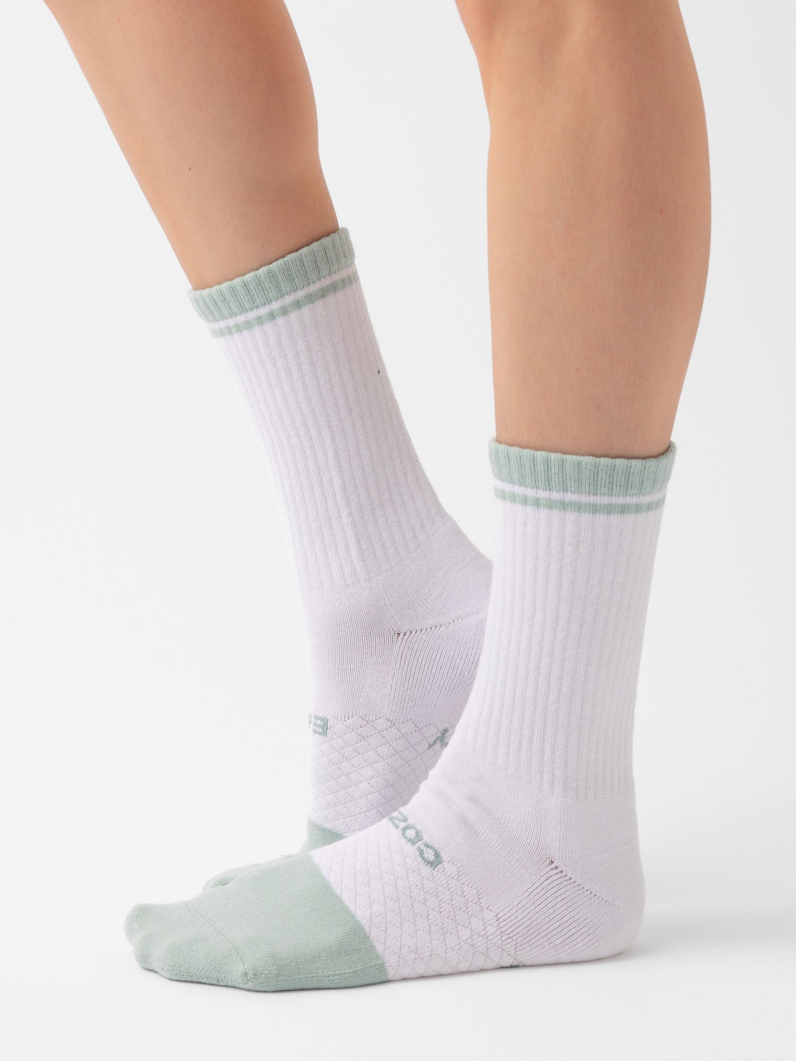 A close-up of a person wearing the "Essential Calf Sock 2-Pack" from Cozy Earth, featuring white crew socks with light green toes, heels, and a green stripe at the top. The mid-calf length socks have a ribbed texture. The person is standing against a plain white background. 