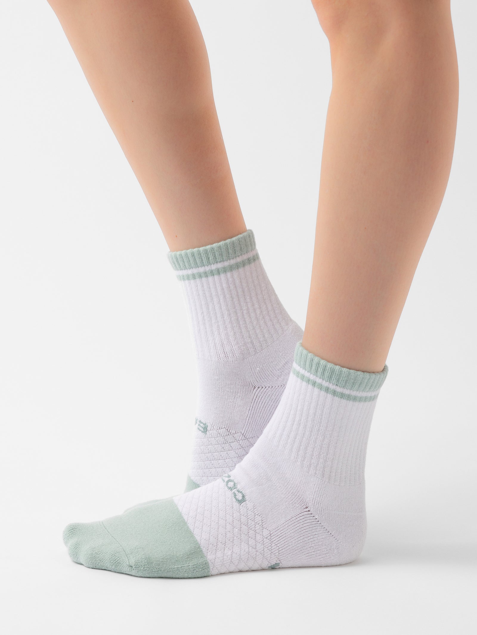 A person is wearing a pair of Cozy Earth's Essential Quarter Socks from the 2-Pack collection, featuring a white base with light green accents on the toes, heels, and cuffs. The socks are designed with a ribbed texture around the ankle and an embossed diamond pattern on the instep. The background is plain white. 