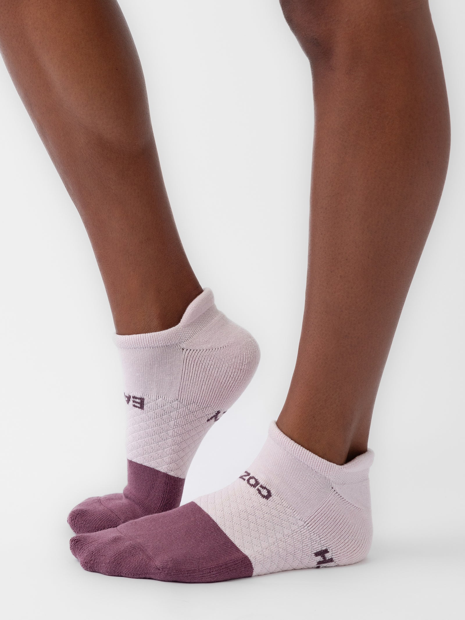 A pair of feet wearing Cozy Earth's Essential Ankle Sock in light pink and purple. These socks feature a darker purple toe and heel, along with a small, patterned texture on the arch. The background is plain white. 