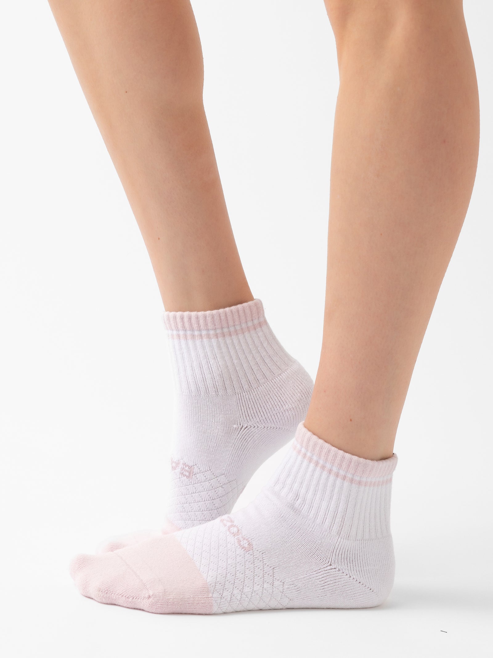 A person wearing white Essential Quarter Socks with light pink accents from the Cozy Earth 4-Pack stands on a plain white background. The socks feature ribbed cuffs and visible stitched patterns along the sides and toes, providing a comfortable and snug fit. 