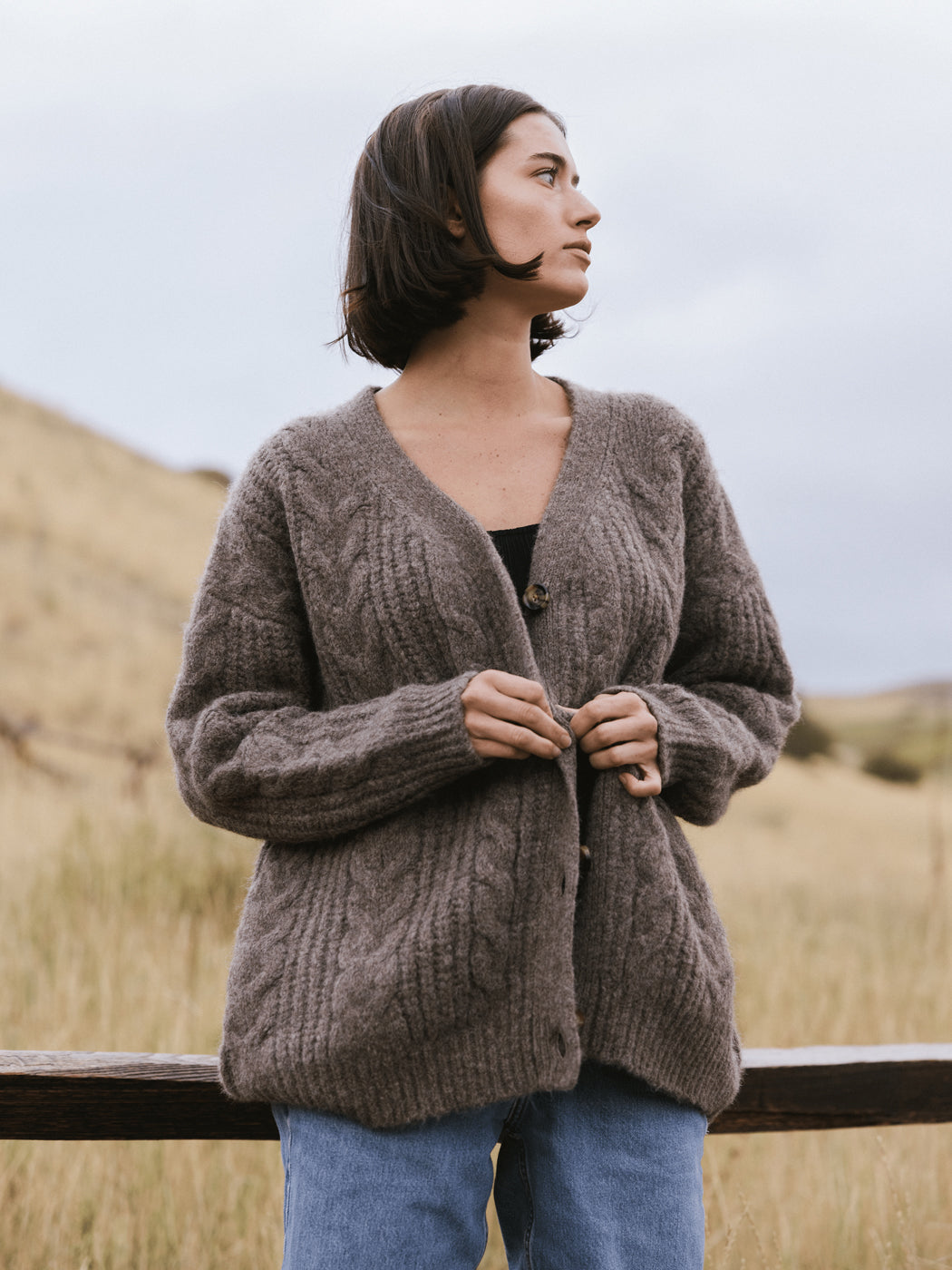 A woman with short dark hair wears the Cozy Earth Oversized Cable Knit Cardigan and blue jeans while standing in a grassy field. She looks to the side calmly; hills and a cloudy sky are softly blurred in the background.