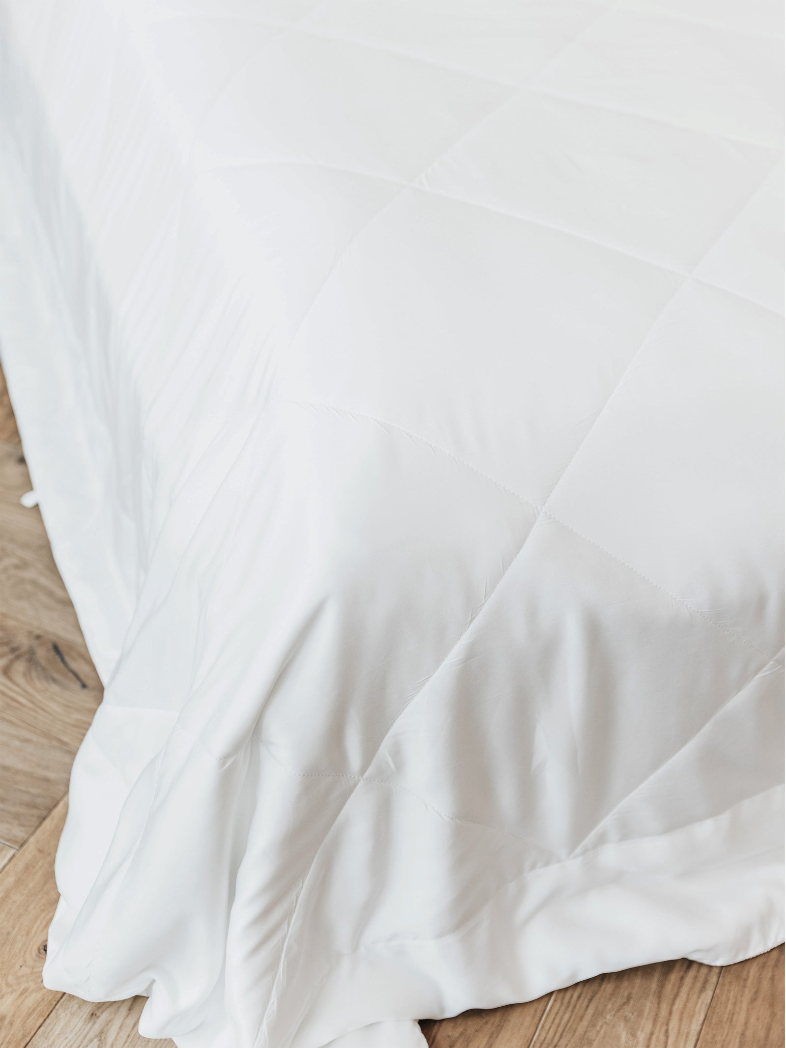 A neatly made bed features the Cozy Earth Bamboo Viscose Comforter, its white quilted surface draped over the edge and resting on a light wooden floor.