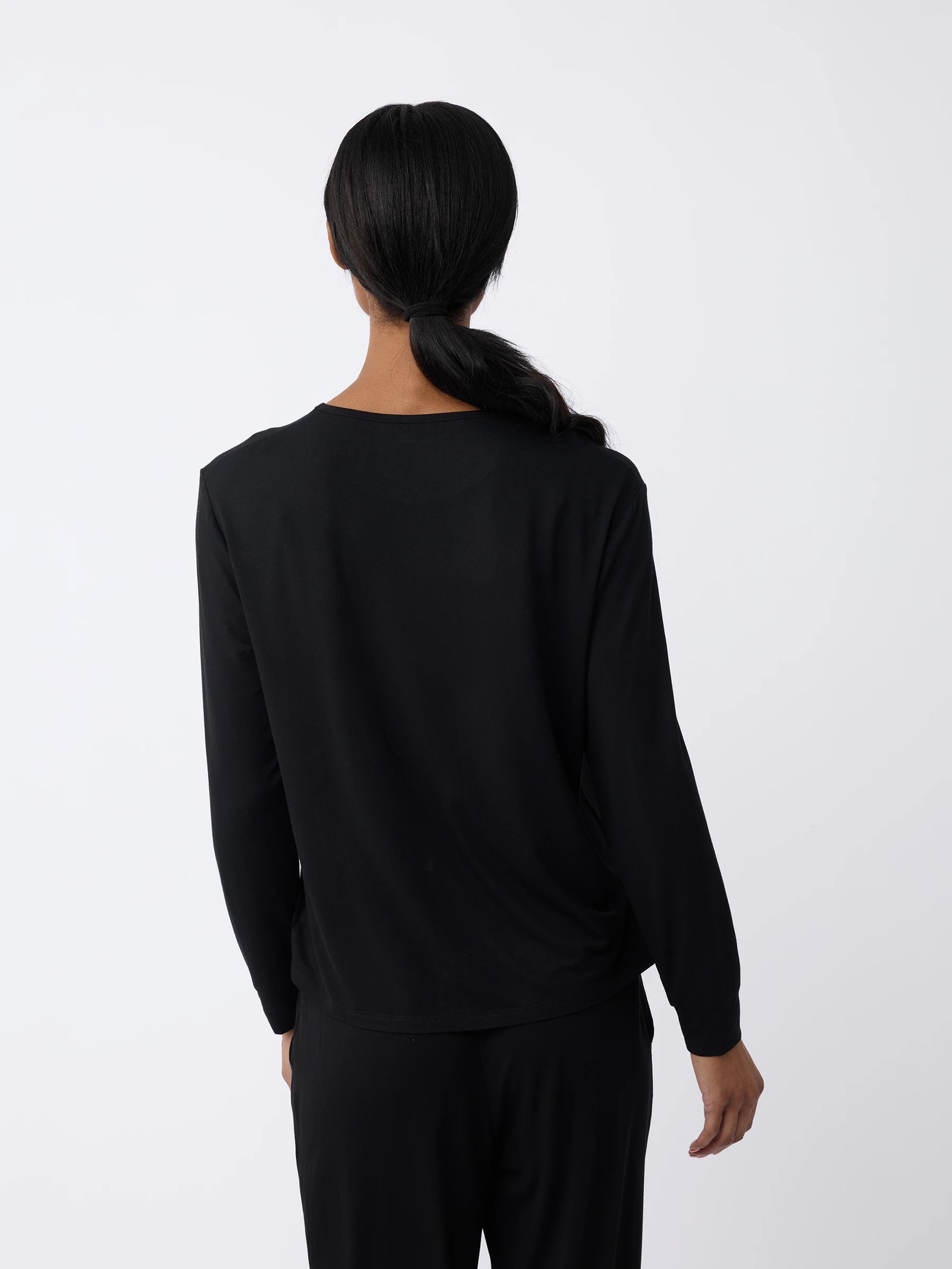 A woman with dark hair in a ponytail is seen from behind, wearing Cozy Earth's Women's Stretch Knit Bamboo Long Sleeve Lounge Tee and black pants against a plain white background. 