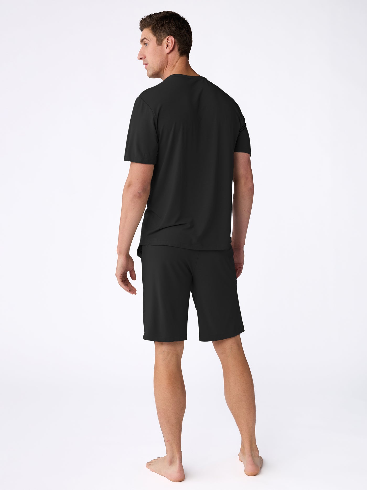 A barefoot man stands facing away, wearing Cozy Earth's Men's Bamboo Stretch-Knit Short Sleeve Pajama Set in black against a plain white background. 