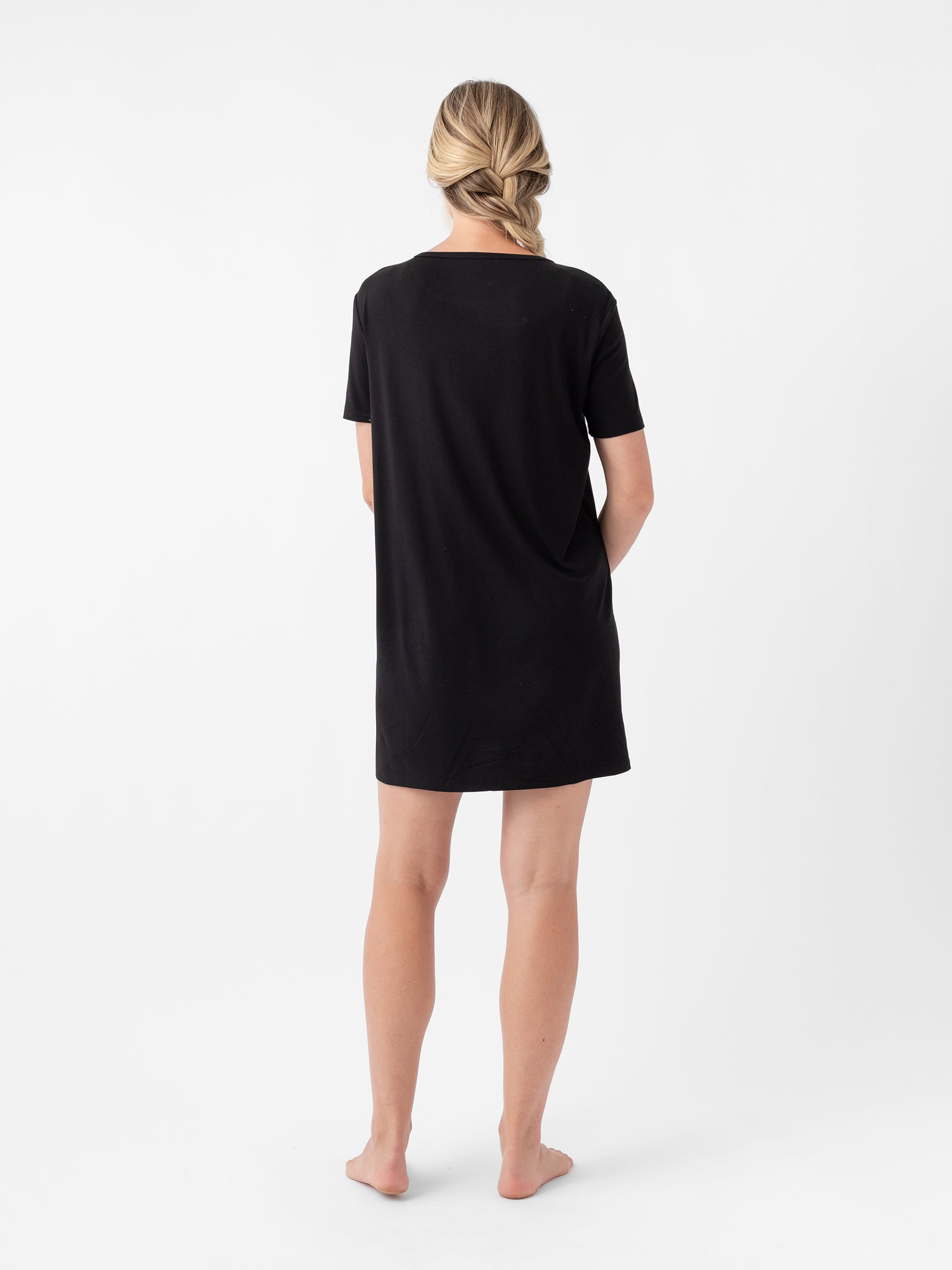 A person with blonde hair is standing barefoot and facing away from the camera. They are wearing a black Cozy Earth Women's Bamboo Stretch Knit Sleep Dress, set against a plain white background. 