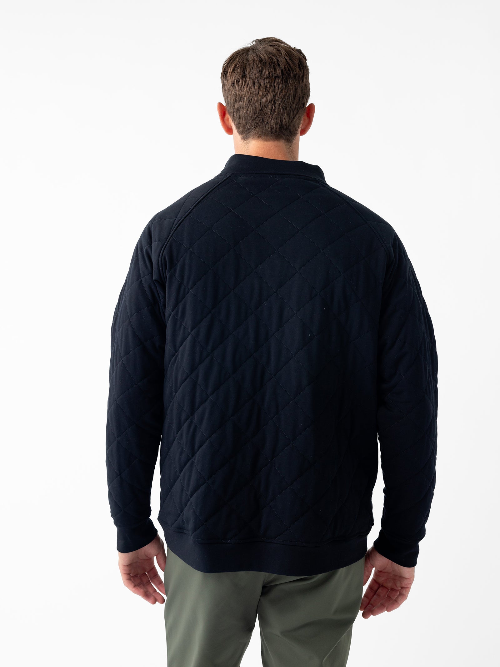 A man with short hair is seen from behind, wearing the Men's Ultra-Soft Bamboo Quilted Snap Pullover by Cozy Earth and olive green pants. The plain white background accentuates the details of the garment. The pullover features a high collar and slightly loose fit with visible stitch patterns. 