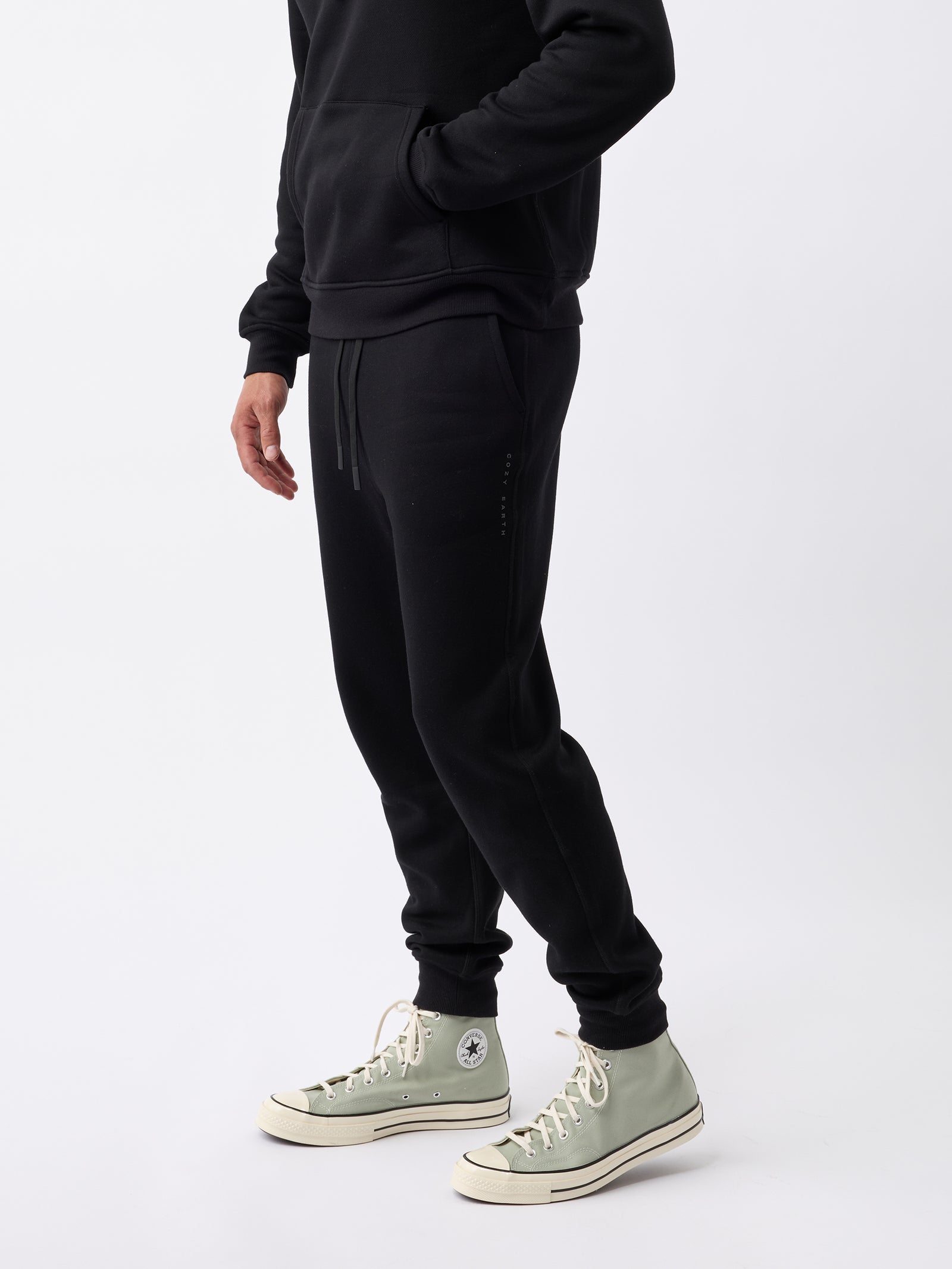 A person wearing Cozy Earth's Men's CityScape Jogger in black and a matching black hoodie stands against a plain white background. They are also sporting light green high-top sneakers, though their face is not visible in the image. 