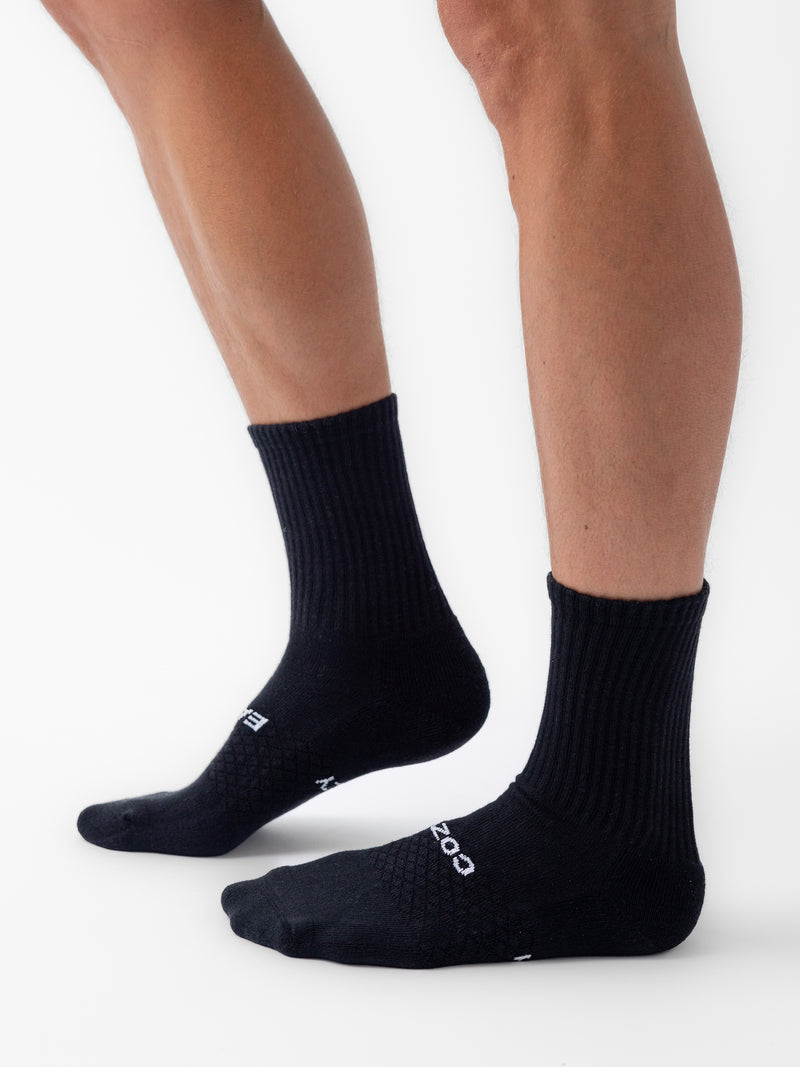 A person wearing Cozy Earth Essential Calf Socks stands on a white background, showing both legs from the knee down. The black socks have a ribbed texture and white text on the top of the foot.