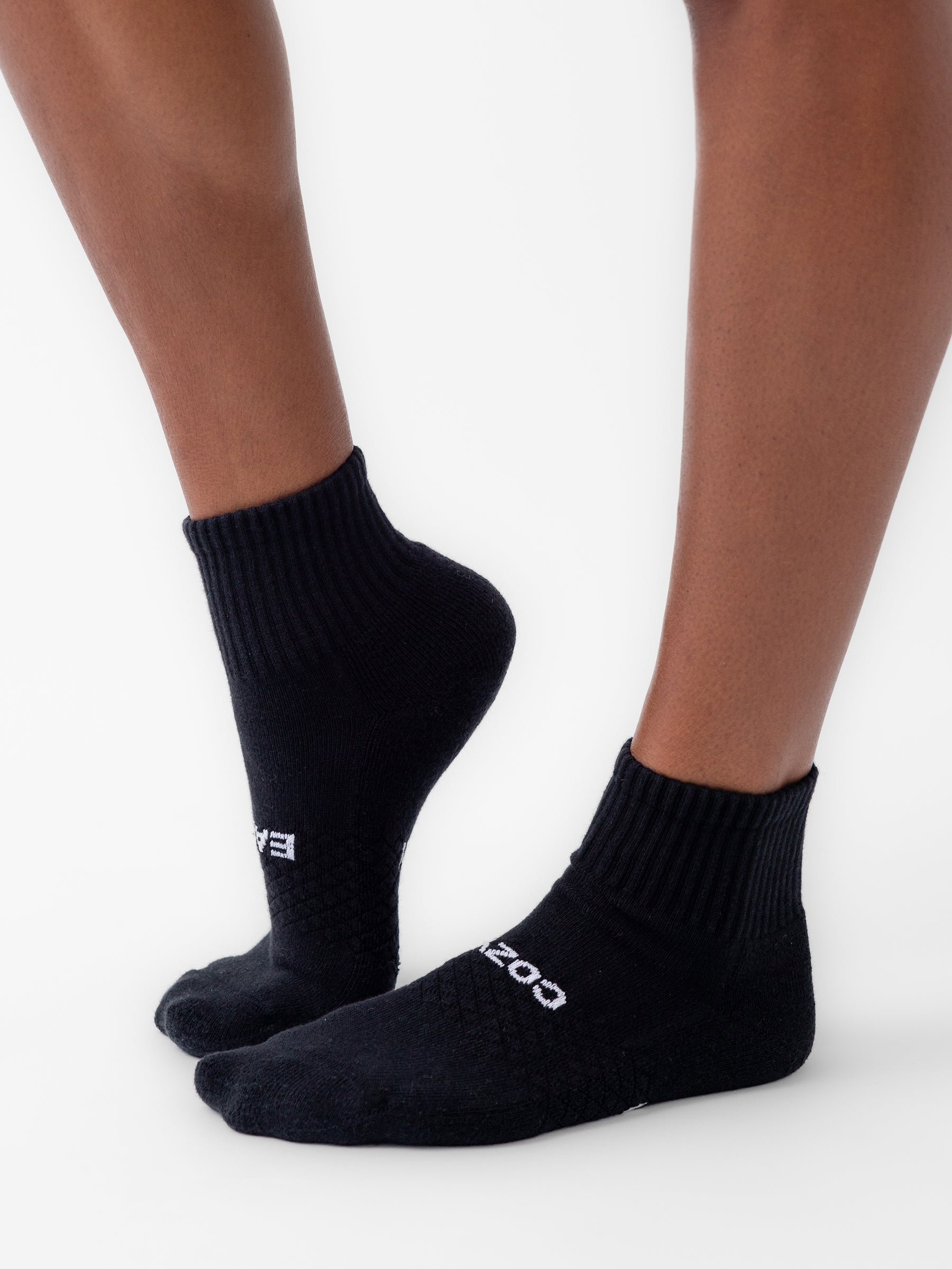 A person wears Cozy Earth's Essential Quarter Sock (6-Pack) in black, featuring a ribbed cuff, textured pattern, and white text near the toes. Only the lower legs and feet are visible as they stand on a white surface. 