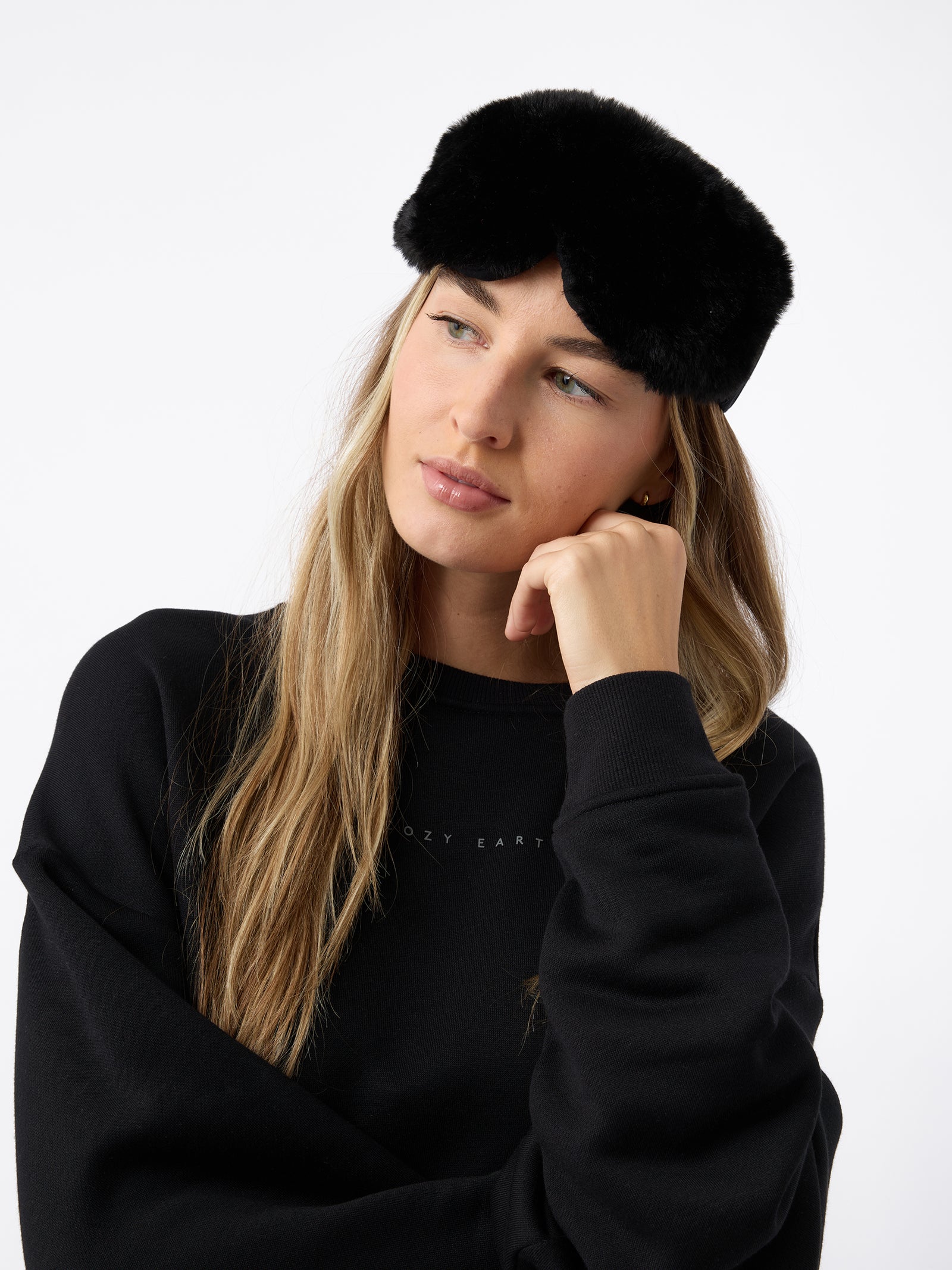 A woman wears the Cozy Earth Faux Fur Sleep Mask in Black. The mask is positioned on her forehead, above her eyes. 