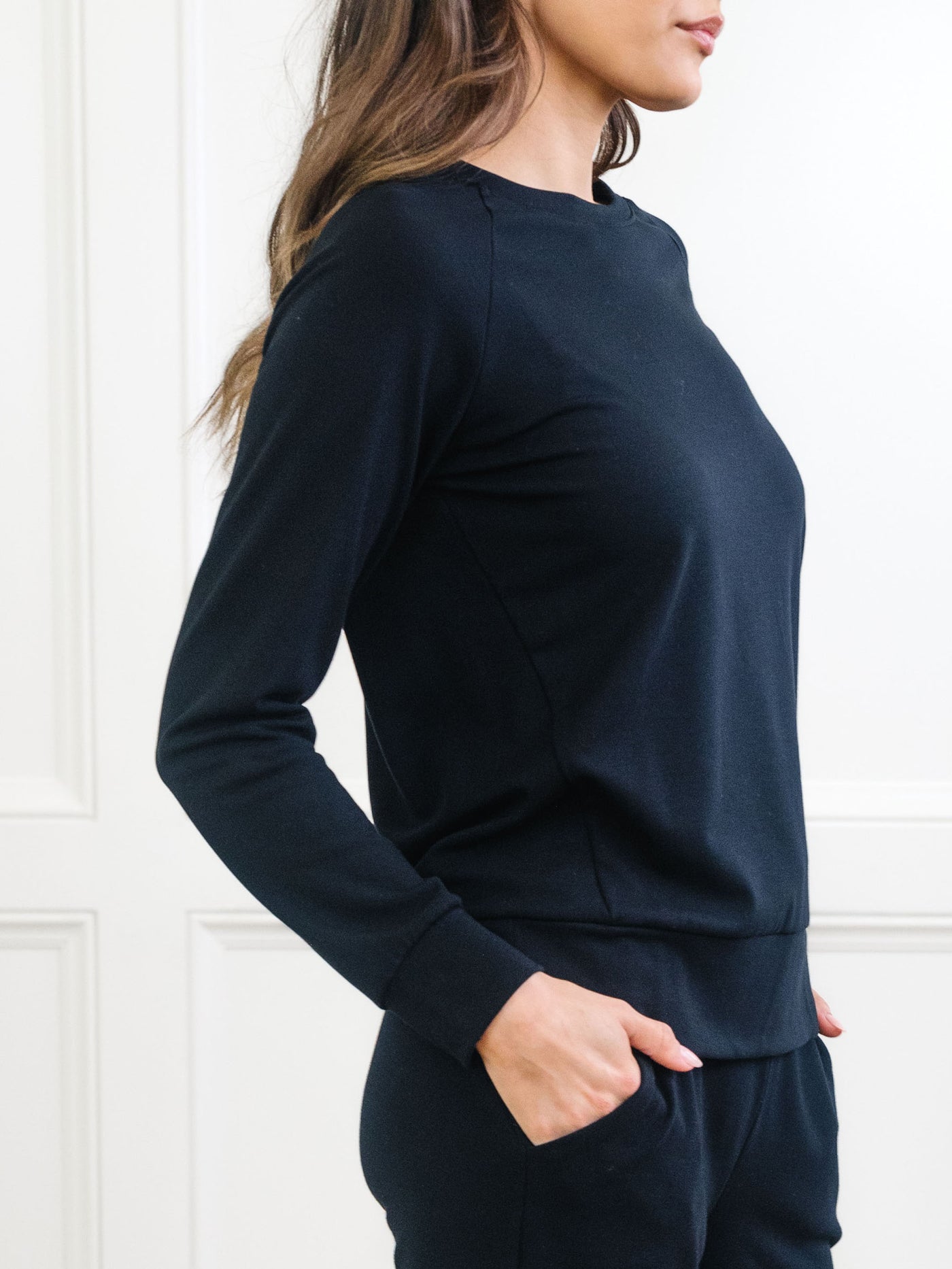 A woman stands sideways in a Cozy Earth Women's Ultra-Soft Bamboo Wide Leg Pull On Pant & Pullover Crew Set with her hair down and one hand in her pocket, against a white-paneled wall. 