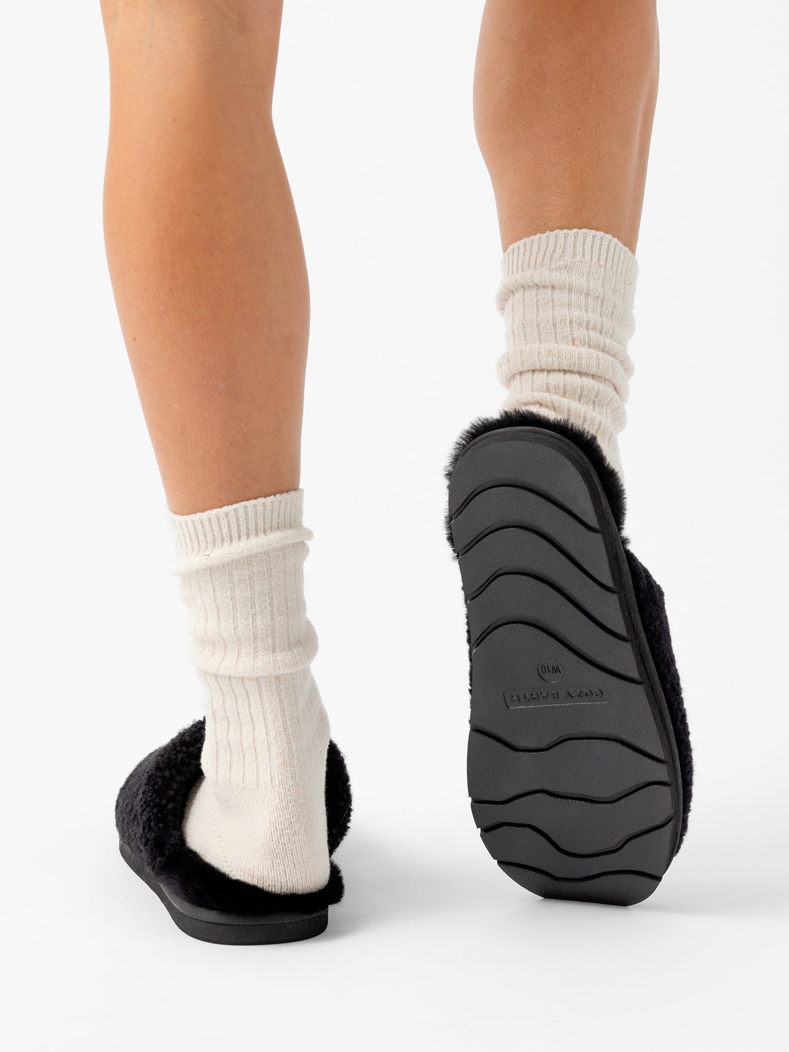 Close-up view of a person walking away, showing their legs from the calf down. They are wearing white socks and Cozy Earth Puffy Sheep Slippers. The slippers have textured rubber soles. The background is plain white. 