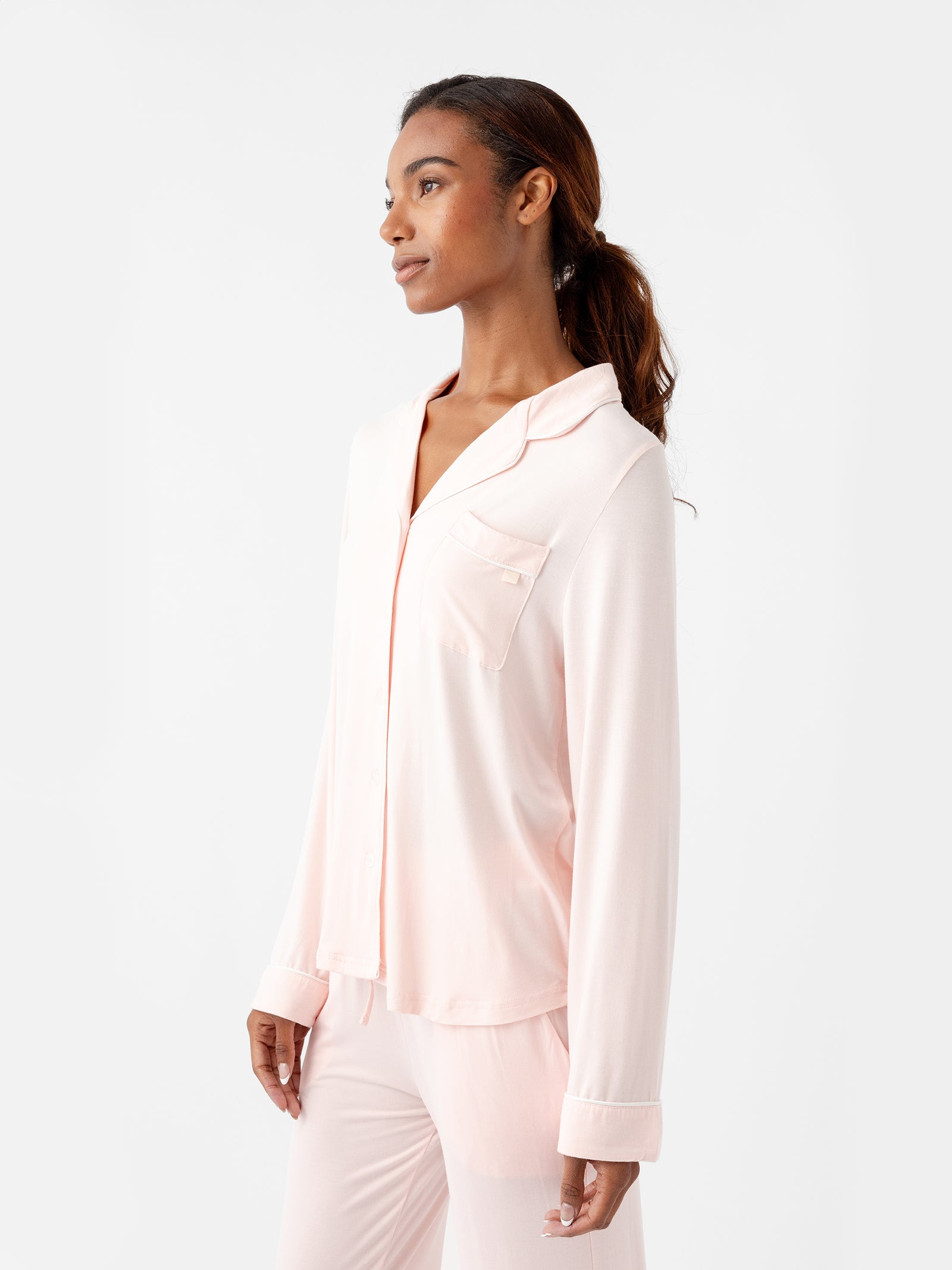 A person with long hair is wearing the Women's Long Sleeve Bamboo Pajama Top in Stretch-Knit TALL by HIDE, featuring a button-up shirt and pants in light pink. They are standing against a plain white background, looking off to the side with a relaxed expression. 