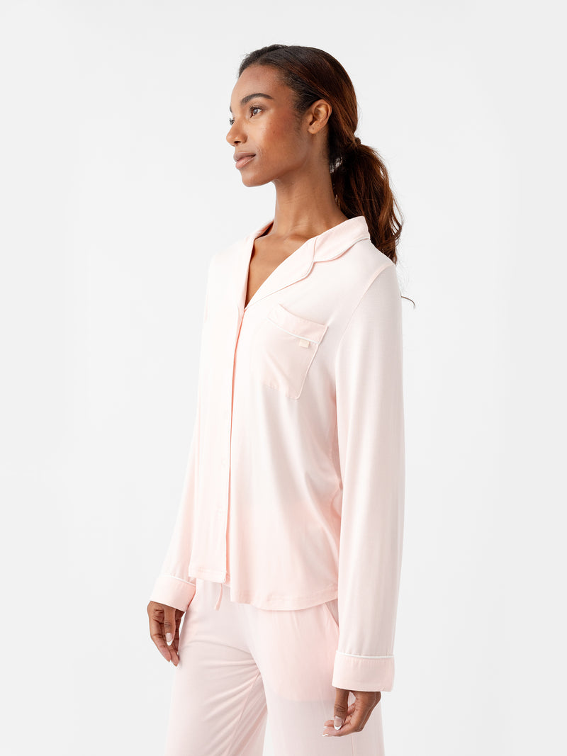 A person with long hair is wearing the Women's Long Sleeve Bamboo Pajama Top in Stretch-Knit TALL by HIDE, featuring a button-up shirt and pants in light pink. They are standing against a plain white background, looking off to the side with a relaxed expression.