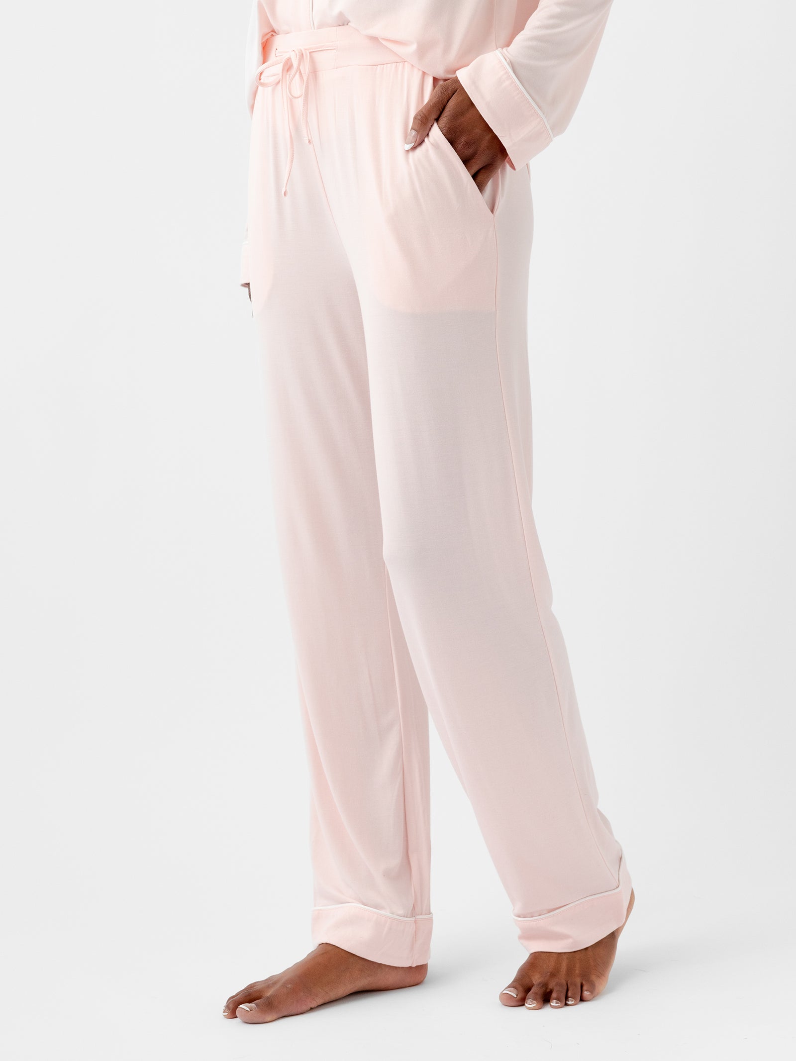 A person stands with their left hand in the pocket of **HIDE's Women's Stretch-Knit Bamboo Pajama Pant TALL** in light pink, which features an elastic waistband with a drawstring. The person is barefoot, and the photo is taken from the thighs down, against a plain white background. 