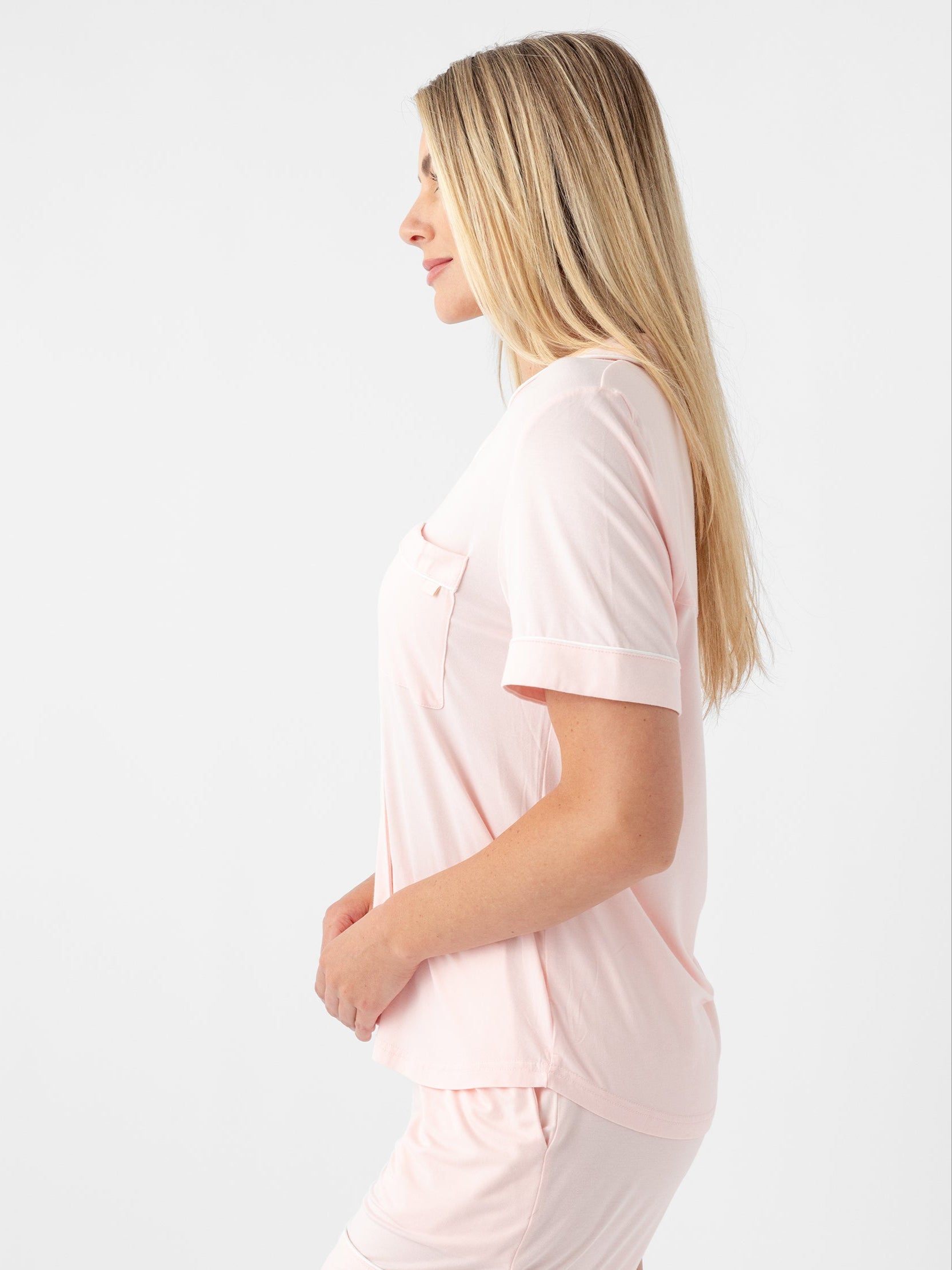 A woman with long blonde hair is standing in profile view, wearing a light pink, short-sleeved Women's Short Sleeve Bamboo Pajama Top in Stretch-Knit by Cozy Earth, paired with matching shorts. The background is plain white. |Color:Blossom