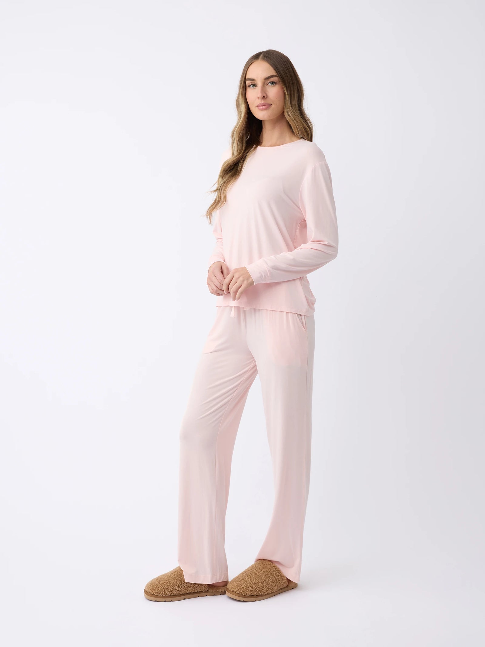 A woman wearing the Cozy Earth Women's Stretch Knit Bamboo Long Sleeve Lounge Tee in light pink with matching pants and brown slippers stands against a plain white background. 