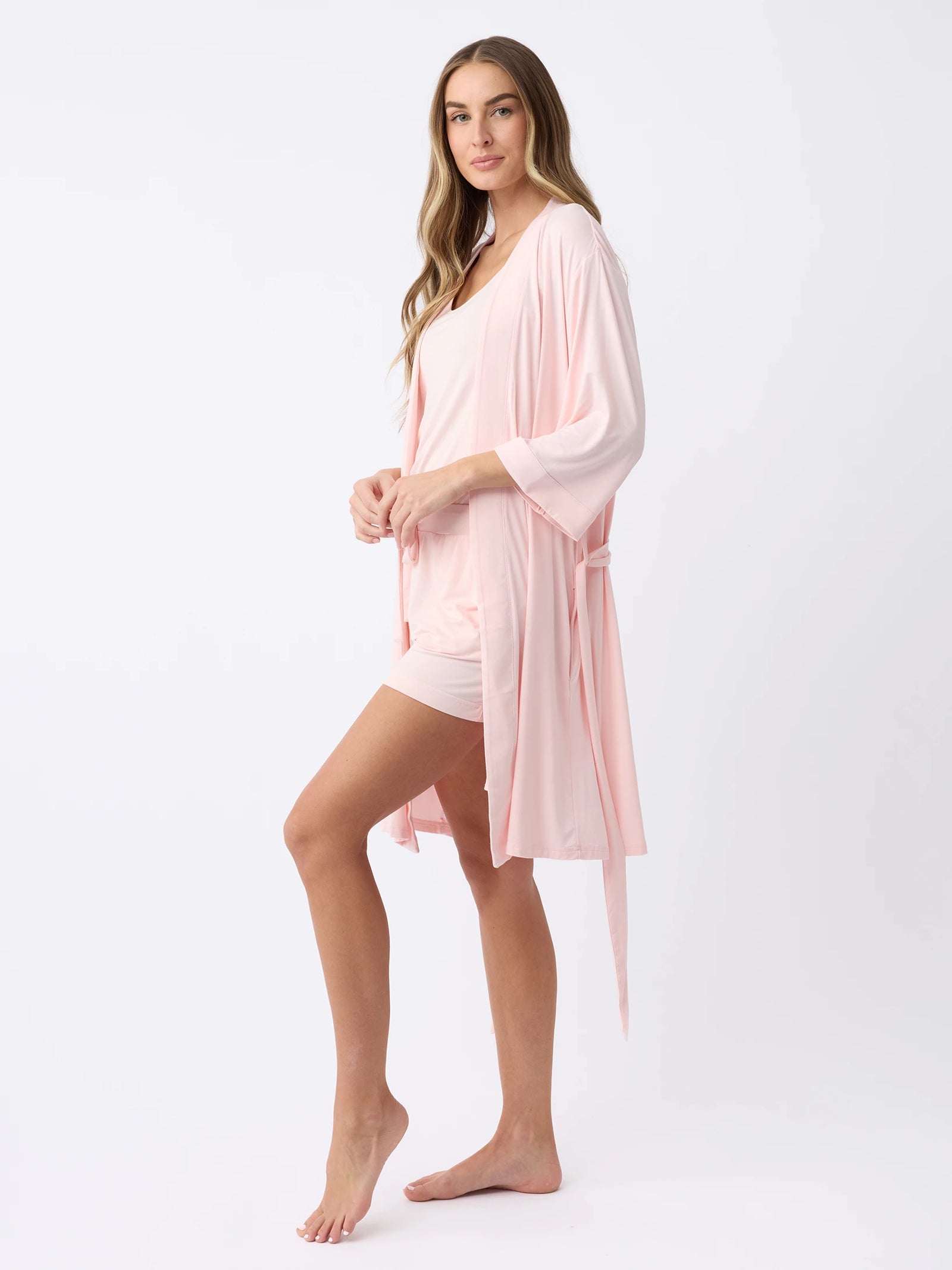 A woman with long, wavy hair stands barefoot in profile against a white background, wearing Cozy Earth's Women's Stretch-Knit Bamboo Kimono Robe in light pink over matching sleepwear. 