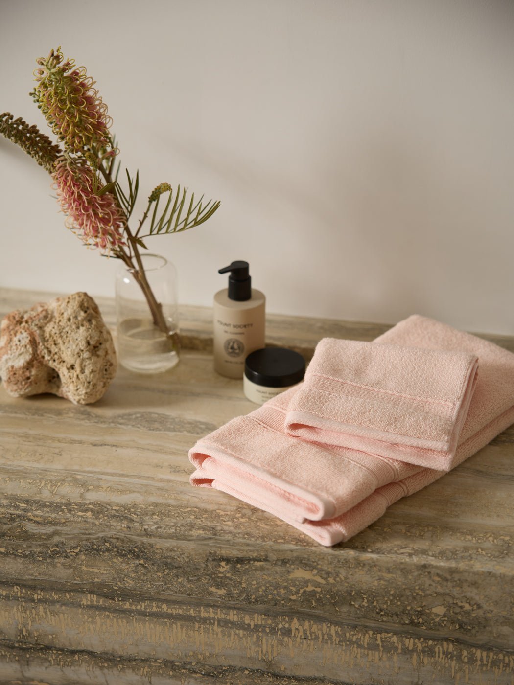 The Complete Luxe Bath Bundle by Cozy Earth features soft pink towels on a marble countertop, complemented by a lotion bottle, small jar, decorative coral, and vase with pink flowers and greenery. 