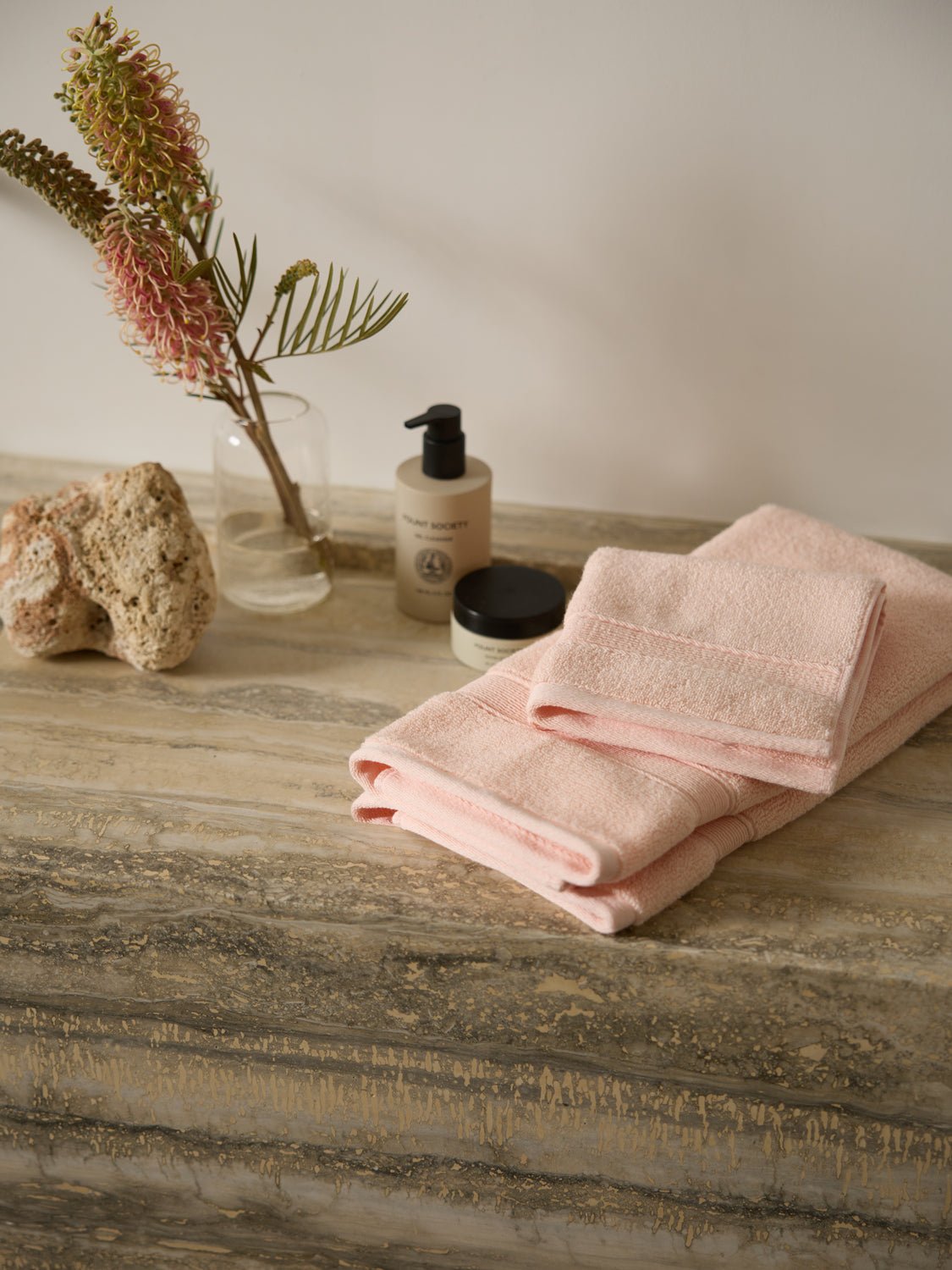 Cozy Earth's Luxe Hand Towels in pink are folded on a marble surface beside a small plant in a glass vase, a large stone, a pump bottle, and a closed jar. The neutral tones and minimalist decor create a soothing atmosphere. 