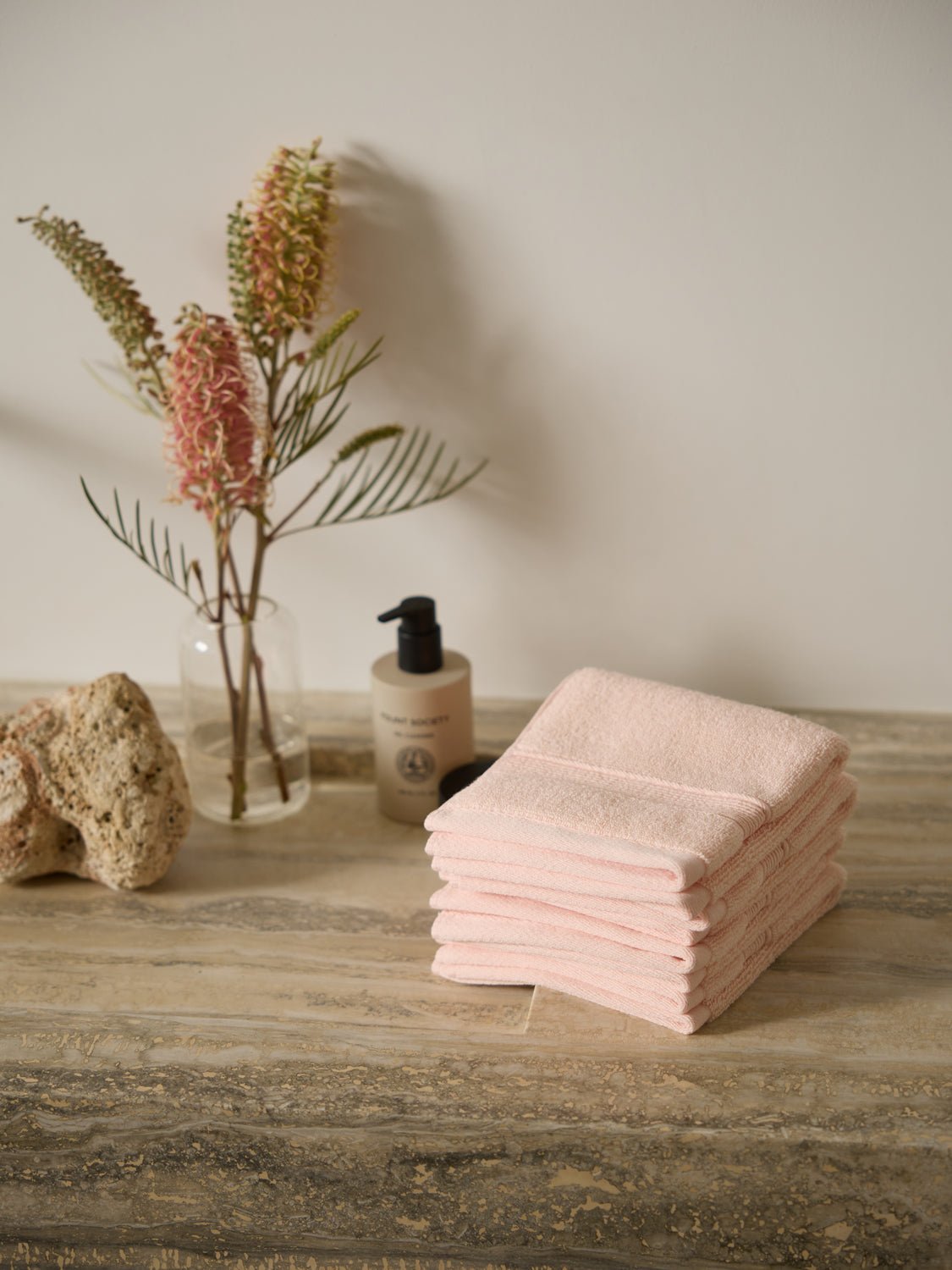 A stack of Cozy Earth Luxe Washcloths in soft pink is neatly arranged on a wooden surface. Nearby are a bottle of liquid soap, a small potted plant with tall flowers, and a piece of coral against an off-white wall. 