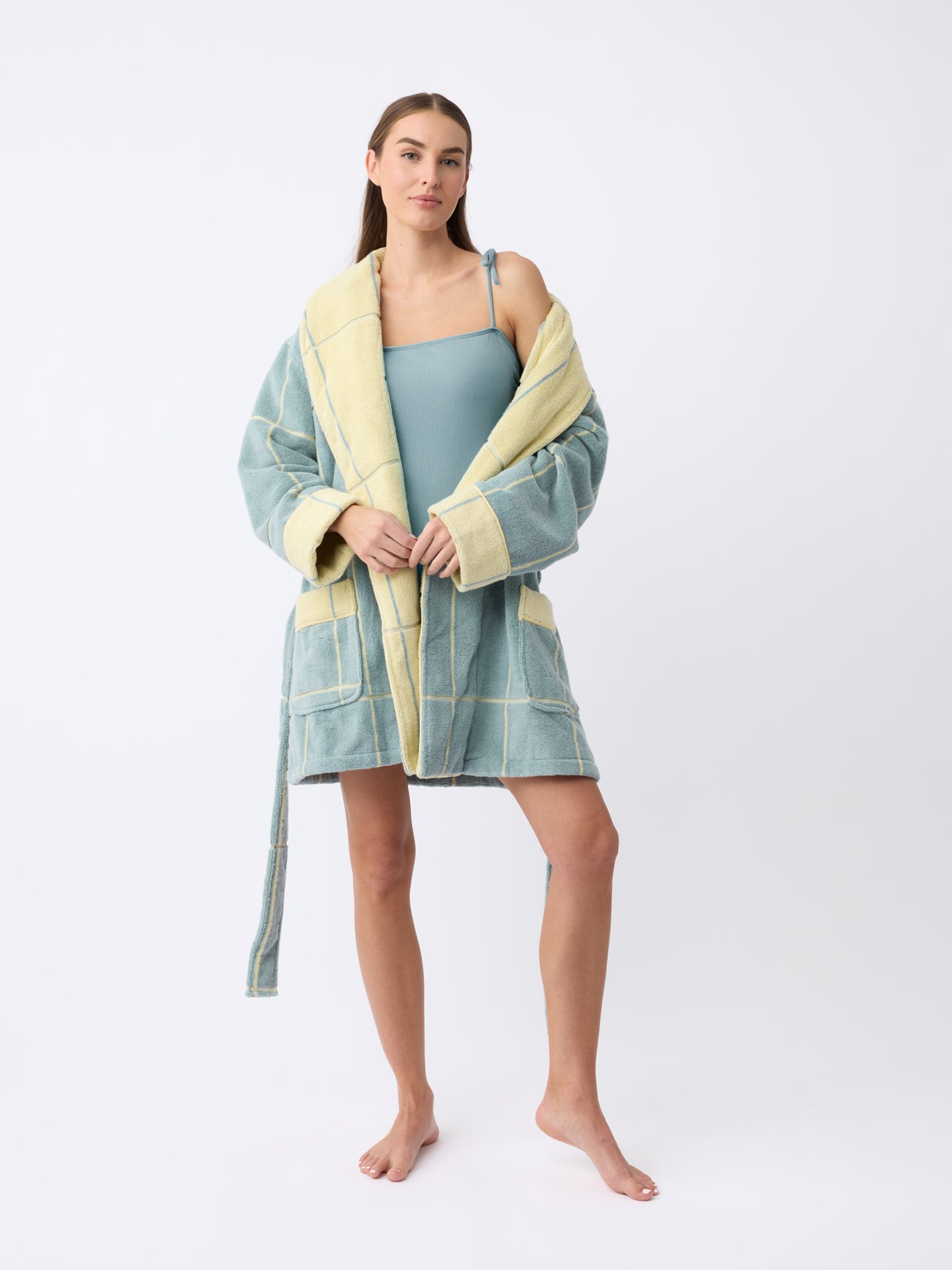 A person stands barefoot against a white backdrop in Cozy Earth's Windowpane Resort Robe, featuring blue and yellow checks over a teal swimsuit, with long brown hair and a confident gaze at the camera. |Color:Blue Lagoon