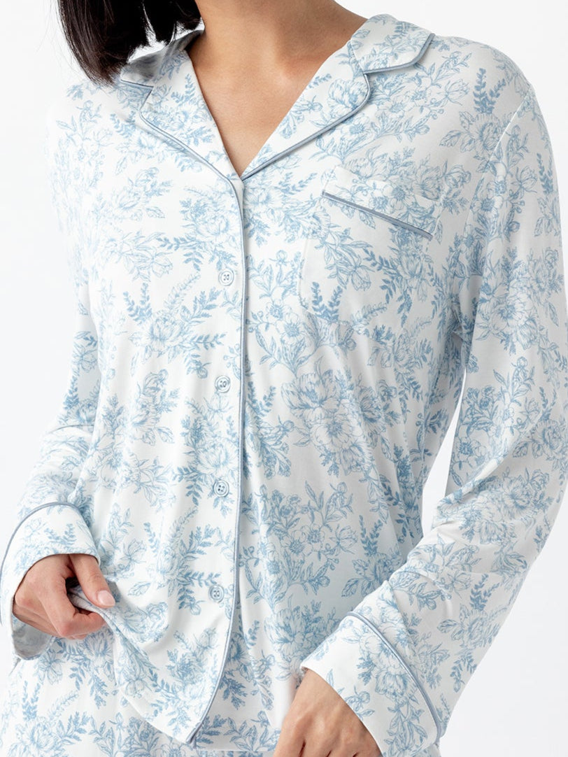 A person with short black hair wears a matching set of Cozy Earth Women's Bamboo Stretch-Knit Long Sleeve Pajama Top in white with a blue floral pattern, standing against a plain white backdrop. They grasp the pajama top with one hand.