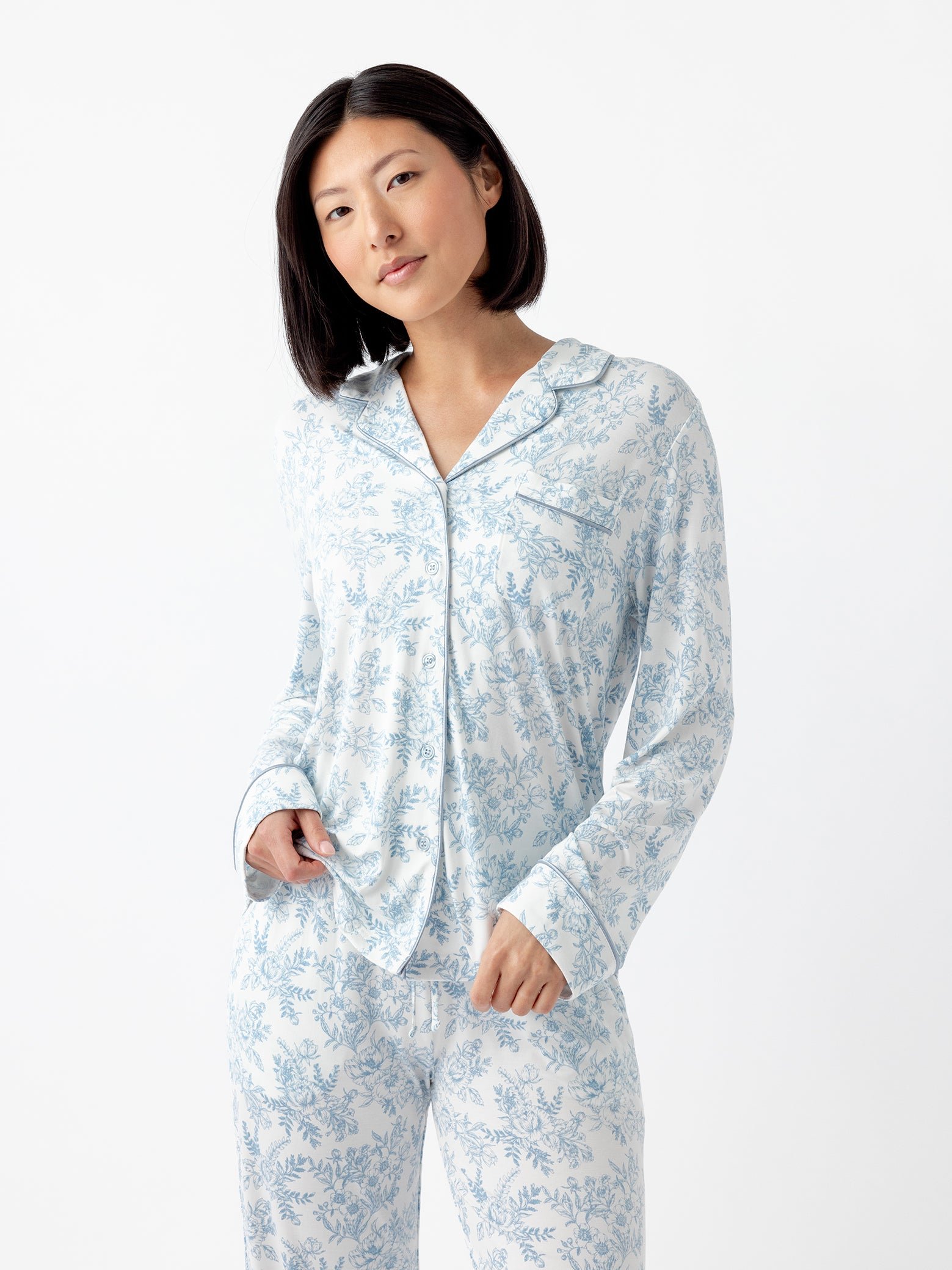 A person with short black hair wears a matching set of Cozy Earth Women's Bamboo Stretch-Knit Long Sleeve Pajama Top in white with a blue floral pattern, standing against a plain white backdrop. They grasp the pajama top with one hand. |Color:Sky Toile