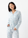 A person with short black hair wears a matching set of Cozy Earth Women's Bamboo Stretch-Knit Long Sleeve Pajama Top in white with a blue floral pattern, standing against a plain white backdrop. They grasp the pajama top with one hand. |Color:Sky Toile