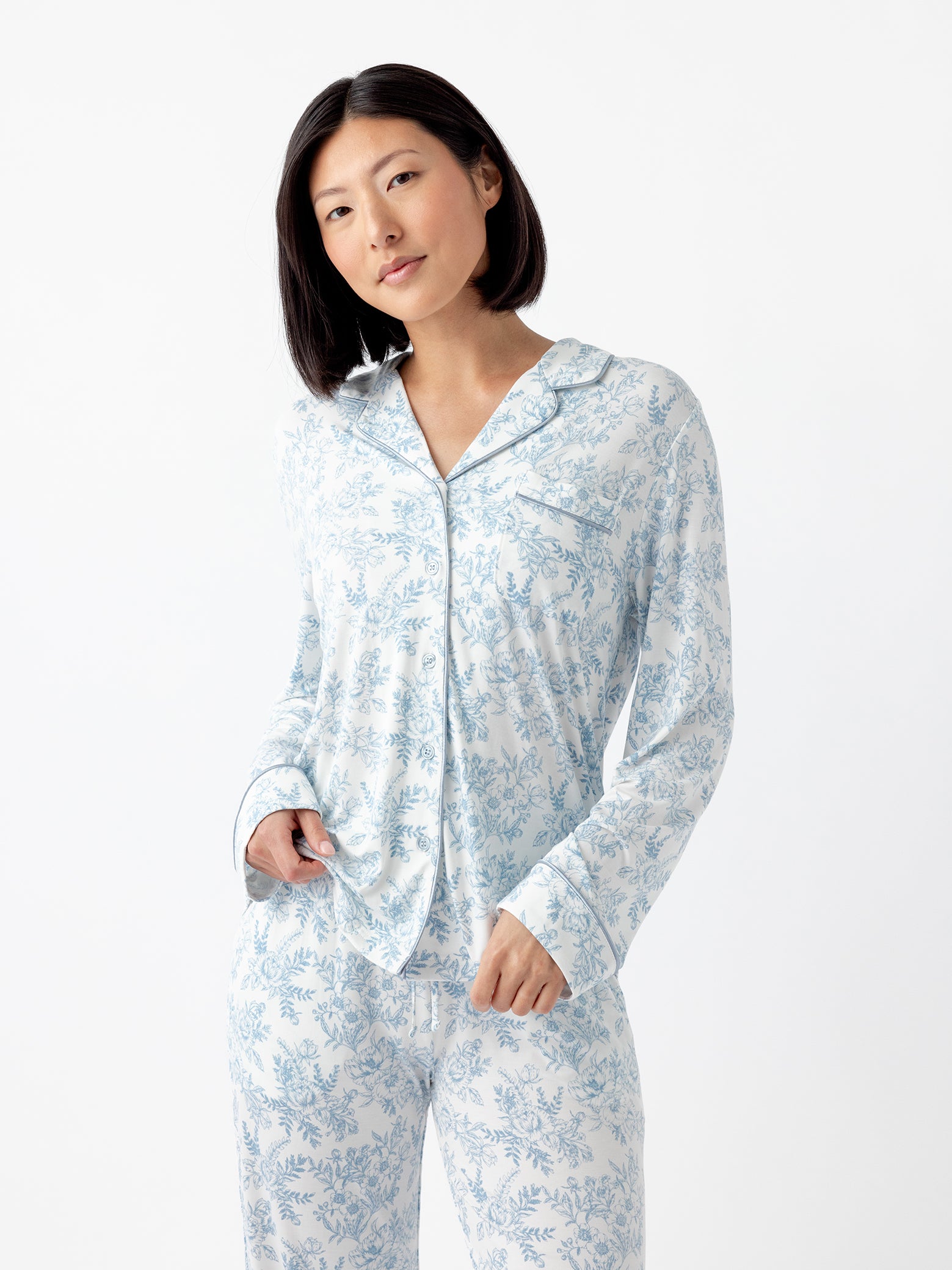 A person with short black hair wears a matching set of Cozy Earth Women's Bamboo Stretch-Knit Long Sleeve Pajama Top in white with a blue floral pattern, standing against a plain white backdrop. They grasp the pajama top with one hand. 