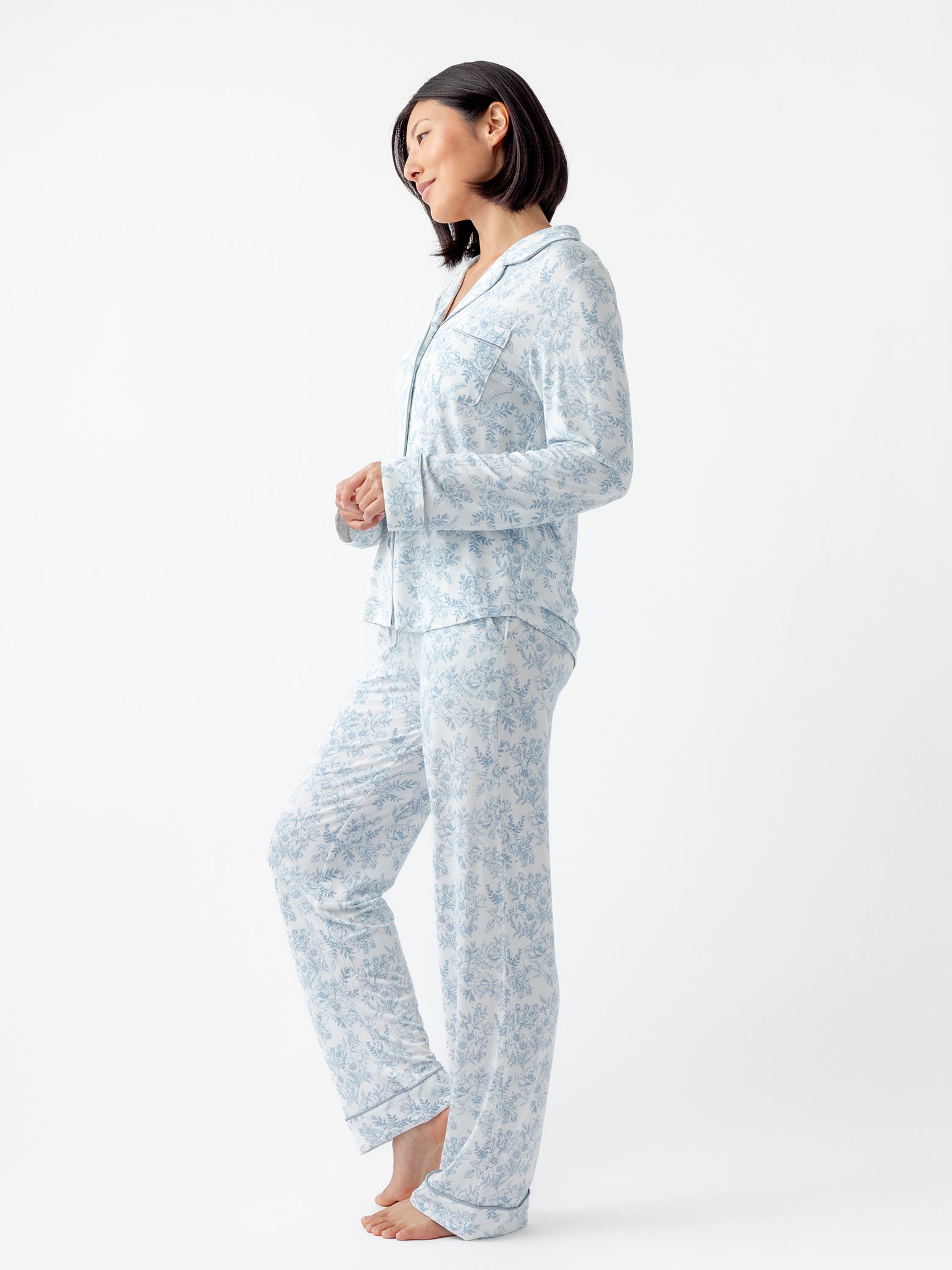 A woman in a Cozy Earth Women's Bamboo Stretch-Knit Pajama Pant in light blue and white floral pattern stands barefoot on a plain white background, looking to the side with a relaxed posture. 