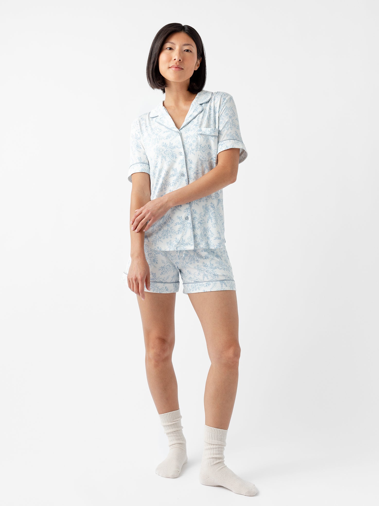 Wearing Cozy Earth Women's Bamboo Stretch-Knit Pajama Short in a light blue floral design and white socks, a person with straight black hair stands against a plain white background, looking slightly to the side with a relaxed expression. 