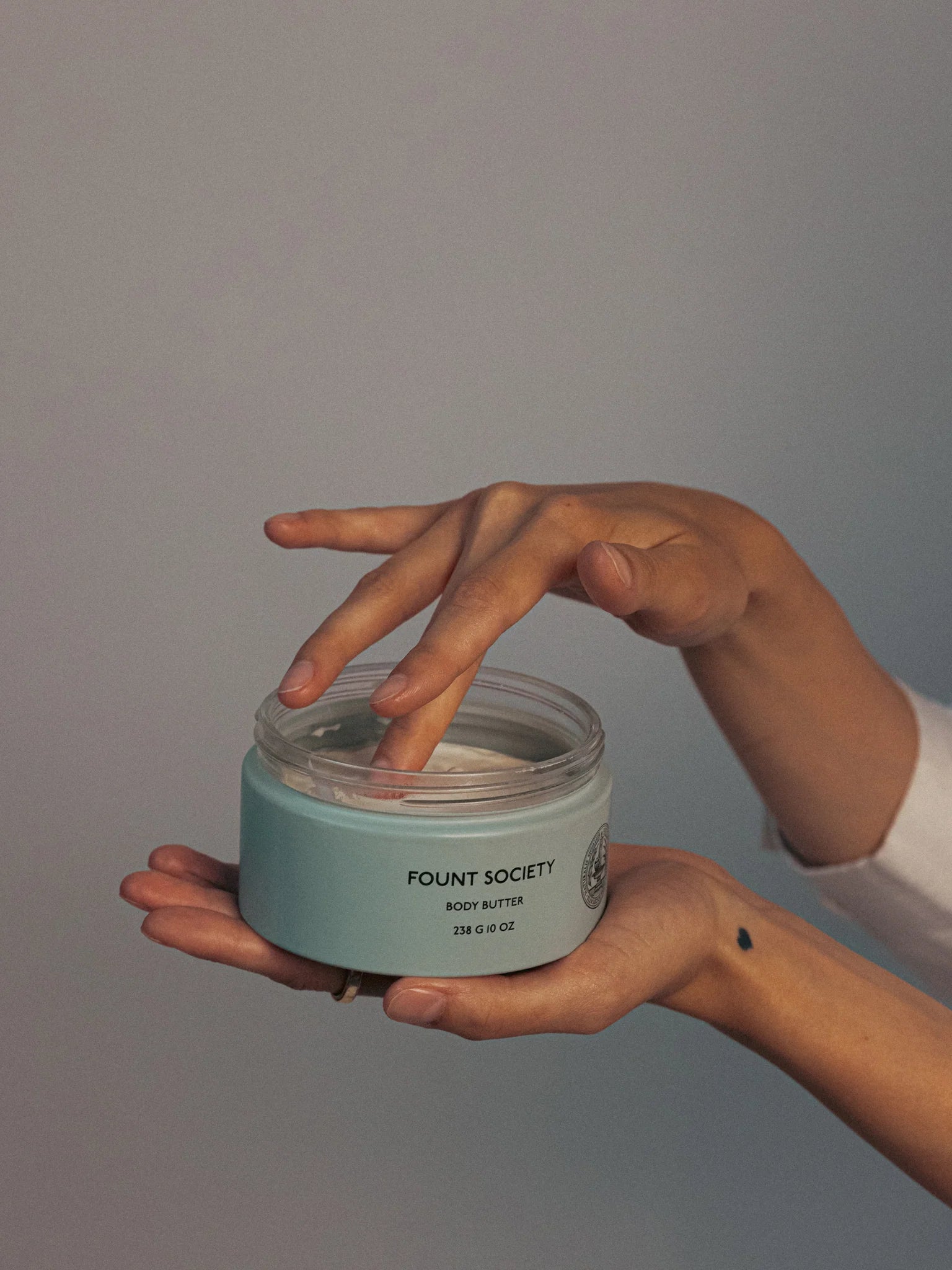 A person holds a jar of Cozy Earth's Bedtime Bundle body butter while dipping a finger into the cream. The teal jar features white text against a soft, neutral background.