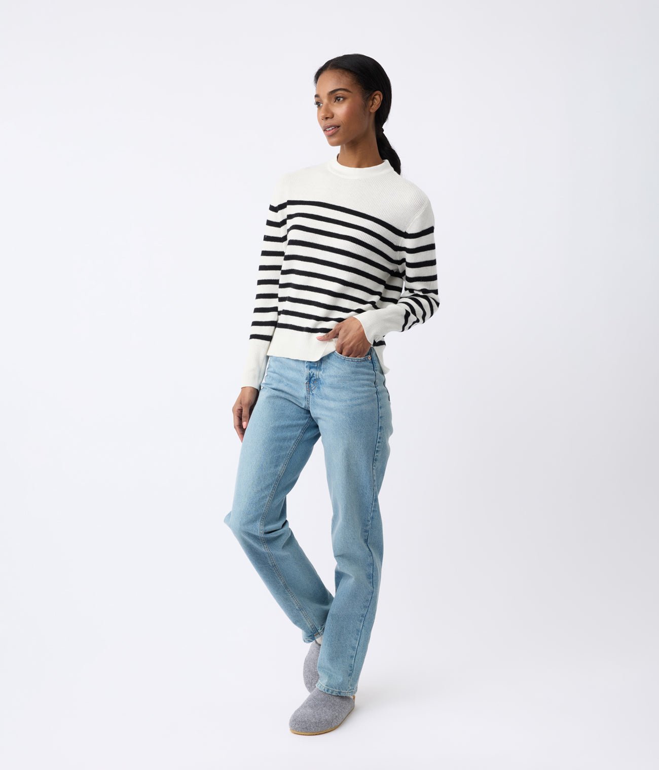 A person in a Cozy Earth Women's Rowan Sweater, blue jeans, and gray shoes stands against a white backdrop, gazing slightly sideways with one hand in their pocket. |Color:Bone/Jet Black Stripe