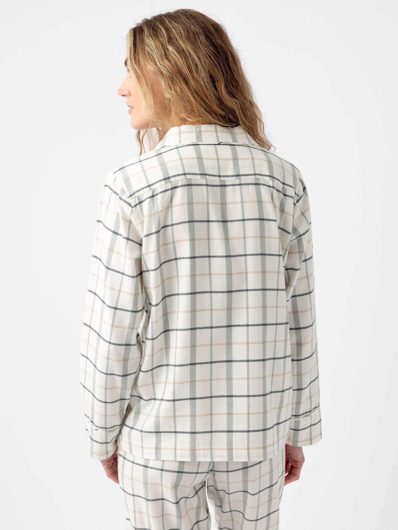 A person with long, wavy blond hair is seen from behind, wearing a Cozy Earth Women's Farmhouse Long Sleeve Pajama Top in a light-colored plaid pattern of wide green and grey checks. The background is plain white. 