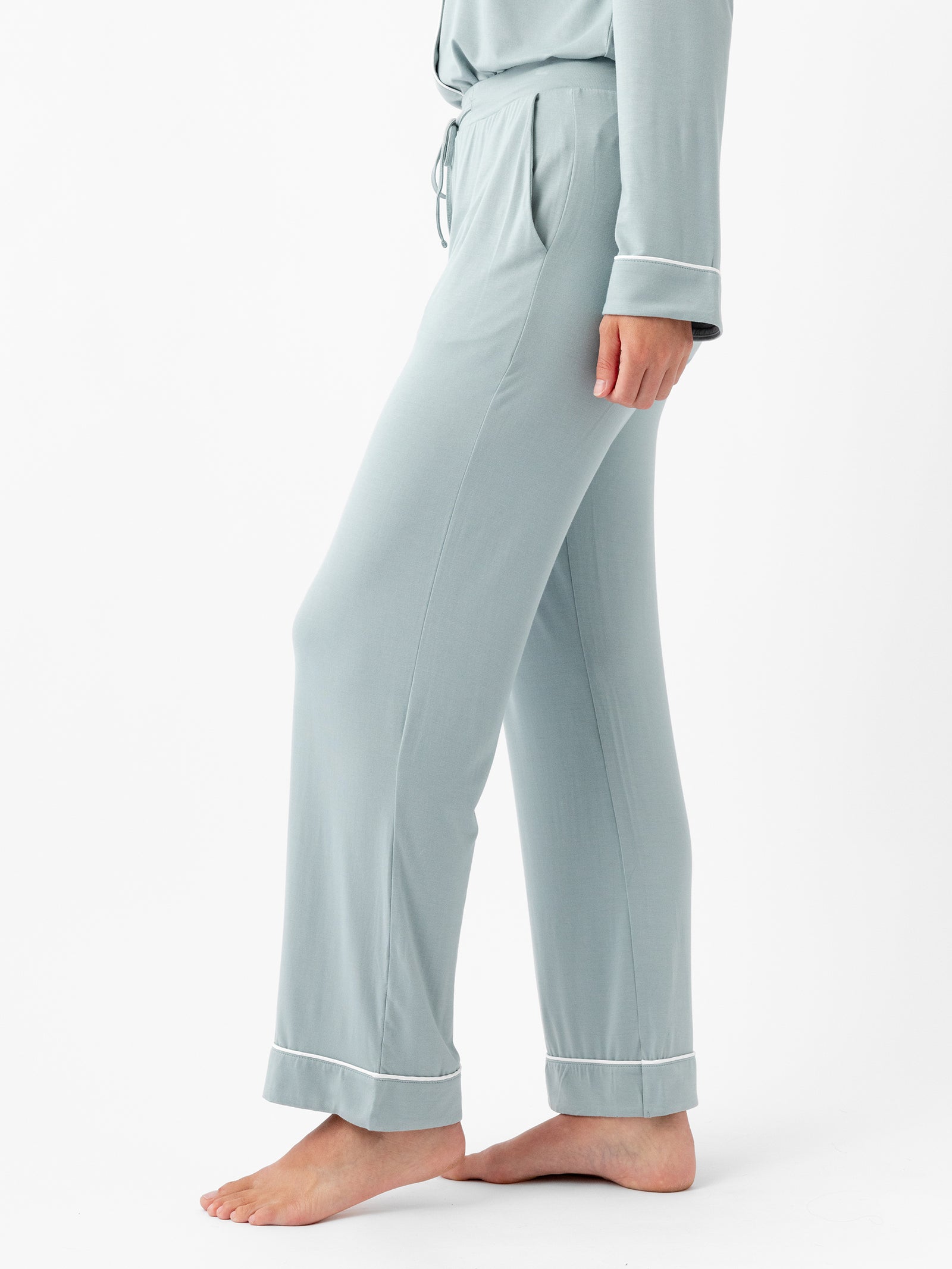 A person is standing sideways facing left, wearing HIDE's Women's Stretch-Knit Bamboo Pajama Pant and a matching top with white trim on the cuffs. The pajama pants are light blue-green, loose-fitting, and ankle-length. The person is barefoot against a plain white background. 