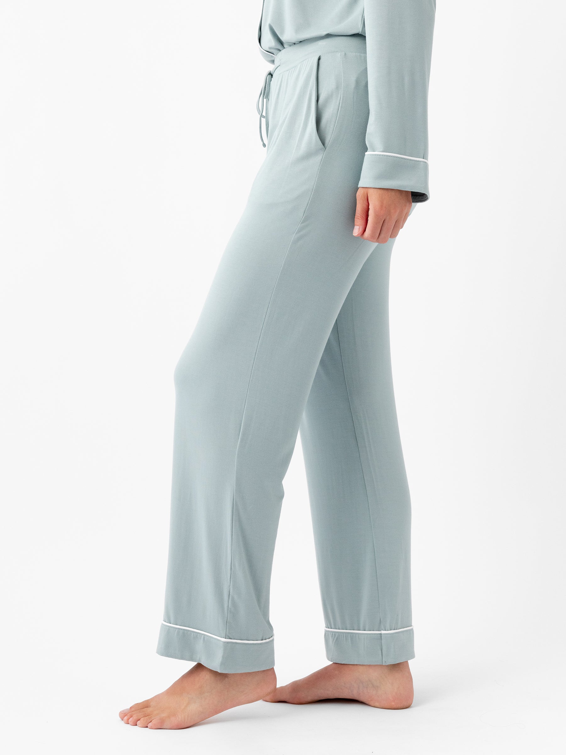 A person is standing sideways facing left, wearing HIDE's Women's Stretch-Knit Bamboo Pajama Pant and a matching top with white trim on the cuffs. The pajama pants are light blue-green, loose-fitting, and ankle-length. The person is barefoot against a plain white background. |Color:Brook