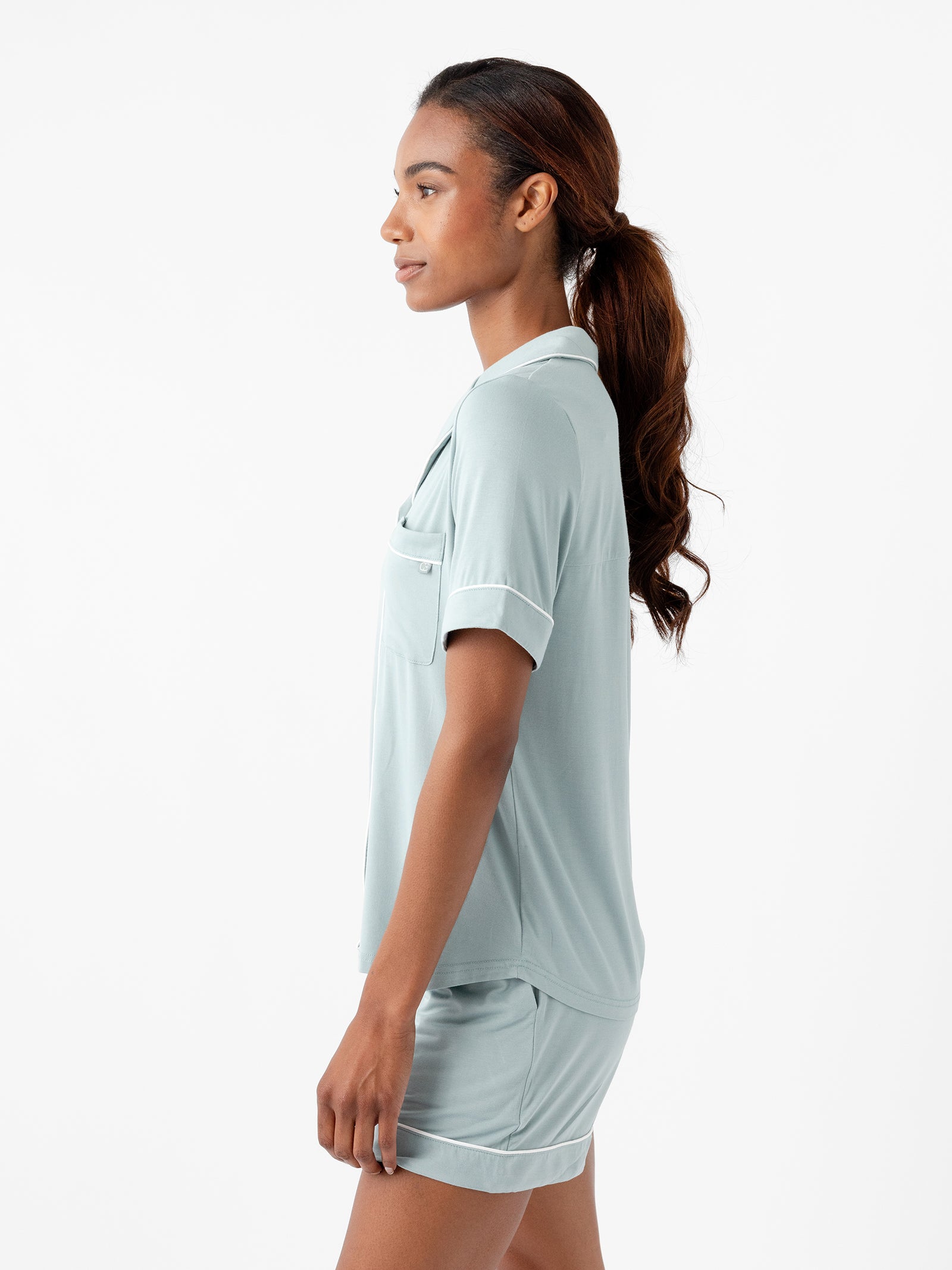 A person with long hair styled in a ponytail is standing in profile view, wearing a Women's Short Sleeve Bamboo Pajama Top in Stretch-Knit and matching shorts from HIDE. The background is plain white. 