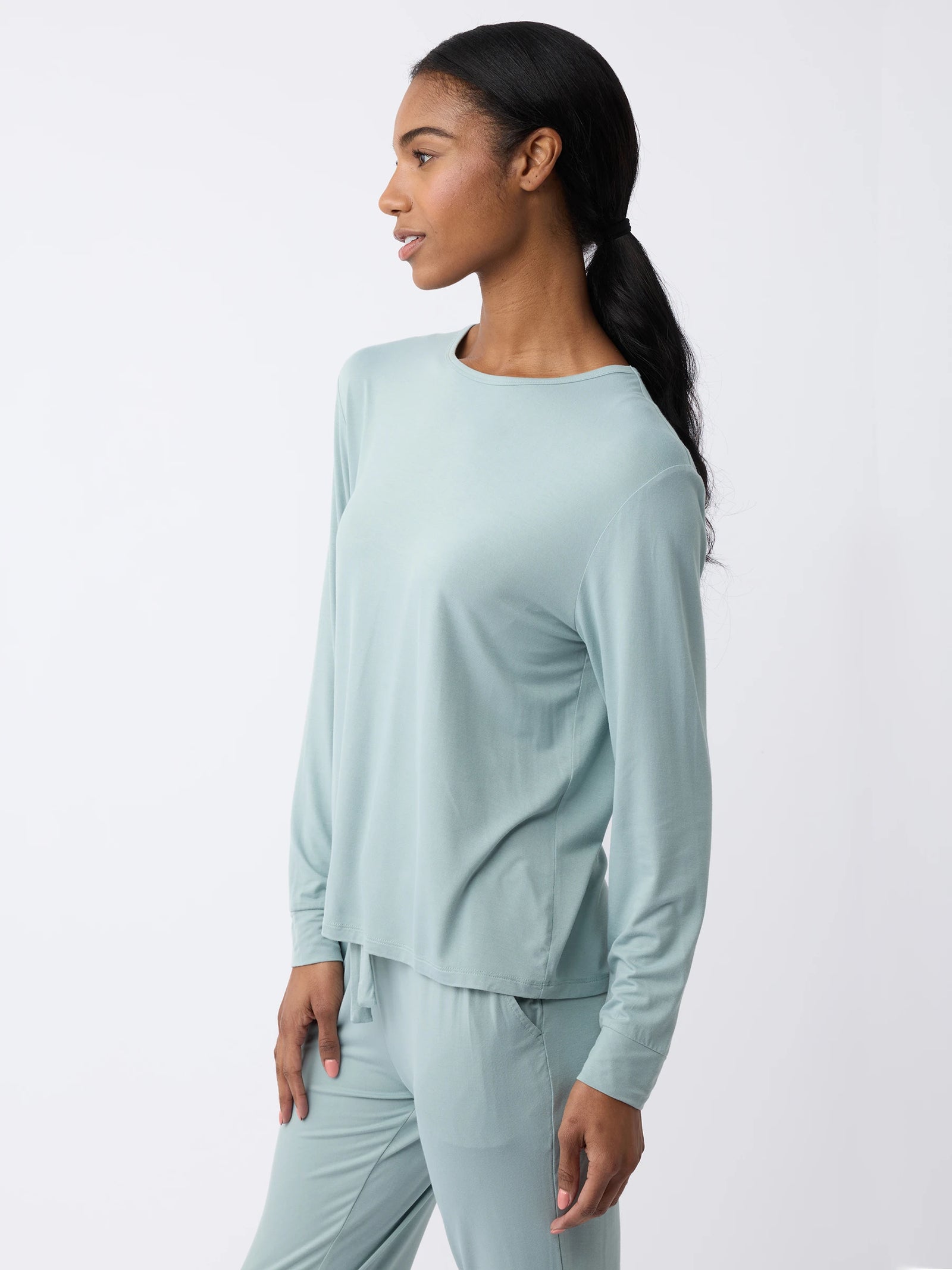 A woman models Cozy Earth's Women's Stretch Knit Bamboo Long Sleeve Lounge Tee in light blue, paired with pants. She stands against a plain white background, looking to the side with a slight smile and her hair tied back in a ponytail. 