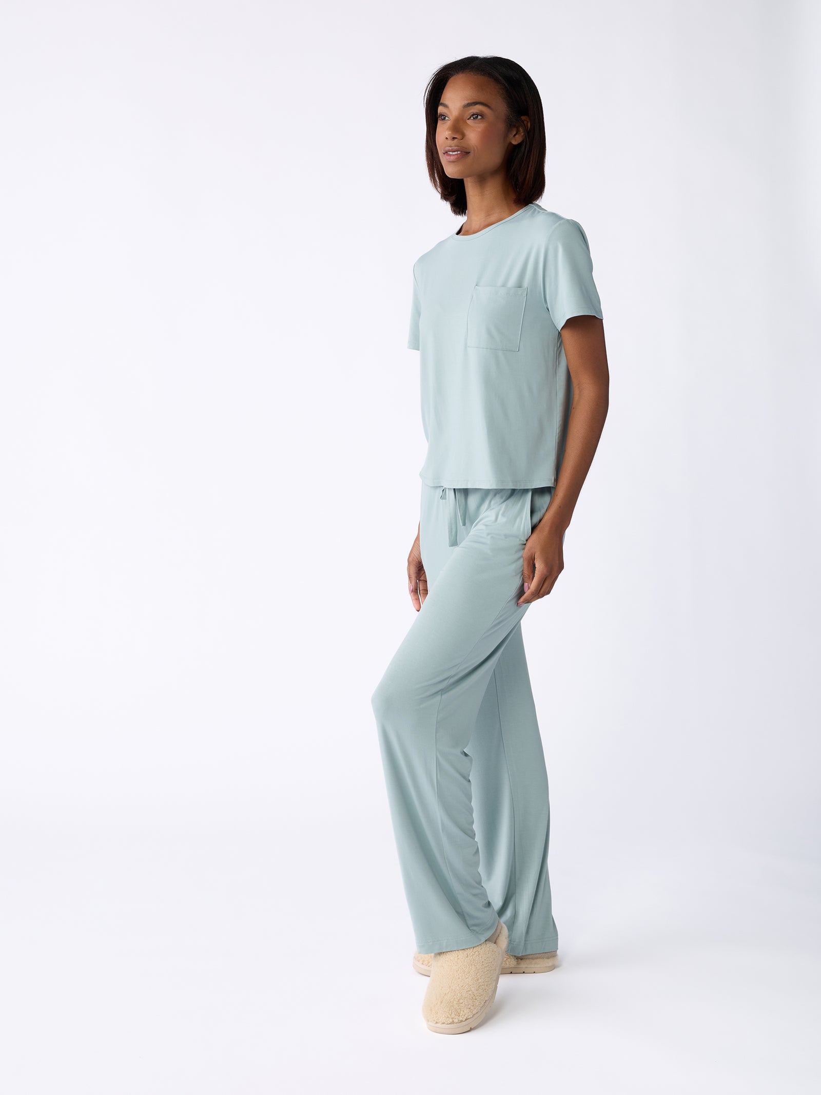 A woman in a light blue Stretch Knit Bamboo Pant from Cozy Earth, paired with beige slippers, stands on a white background. The pajama set features a short-sleeve top with a pocket and loose-fitting pants. She rests her hands by her sides, smiles gently, and glances off to the side. 