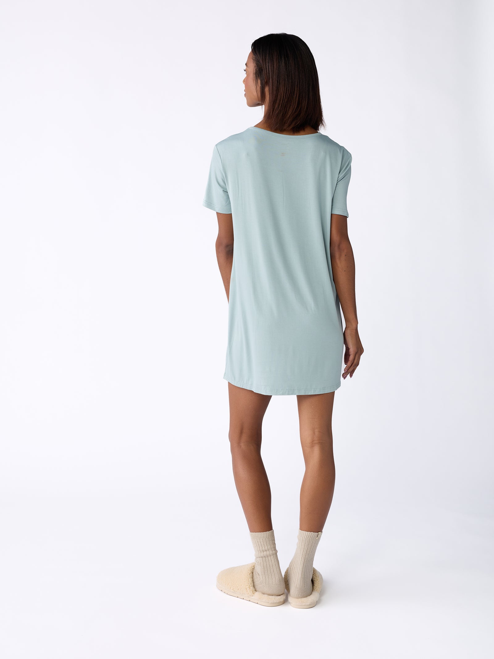 A person is wearing a Women's Bamboo Stretch Knit Sleep Dress by Cozy Earth in light blue, standing with their back to the camera. They have short brown hair and are wearing beige socks and slippers, set against a plain white studio background. 